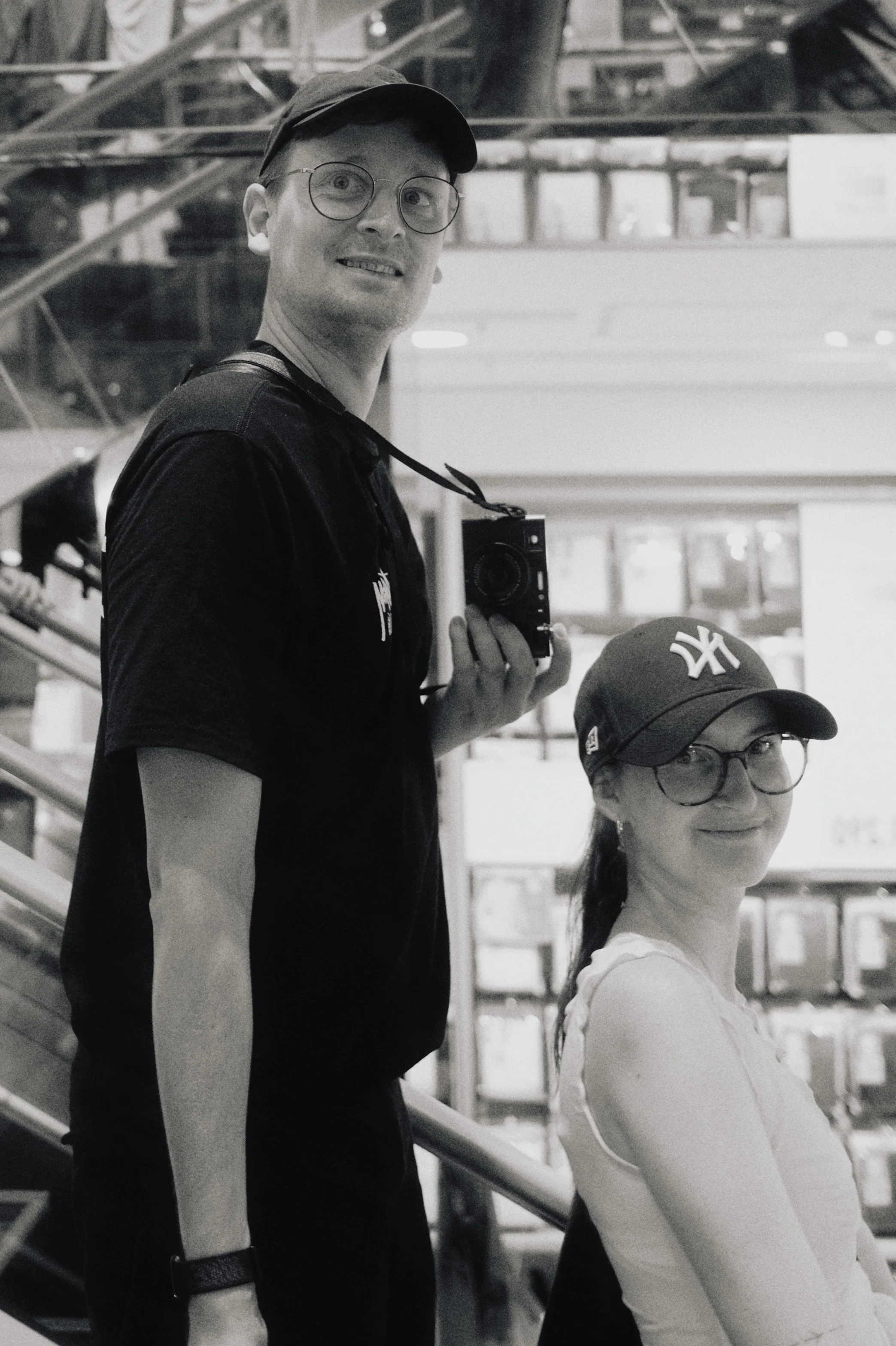 Ein Mann und eine Frau, beide tragen Brillen und Caps, posieren für ein Selfie in einem öffentlichen Ort, möglicherweise eine Bahnstation oder ein Einkaufszentrum, im Schwarz-Weiß-Stil.