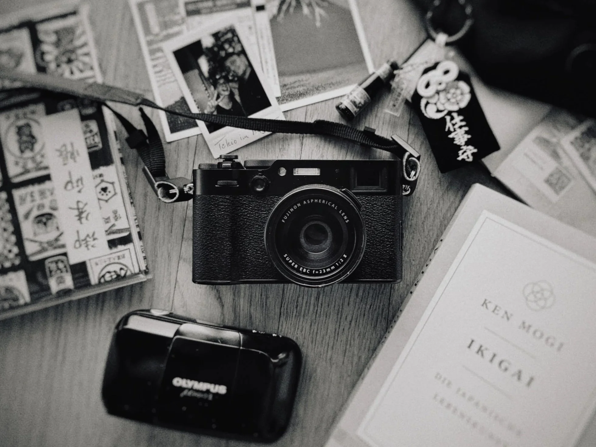 Schwarz-weiße Fotografie von einer Tischszene mit einer Kamera, mehreren Postkarten, einem Buch, einem Olympus-Fotoapparat, Andenken und Zigaretten.
