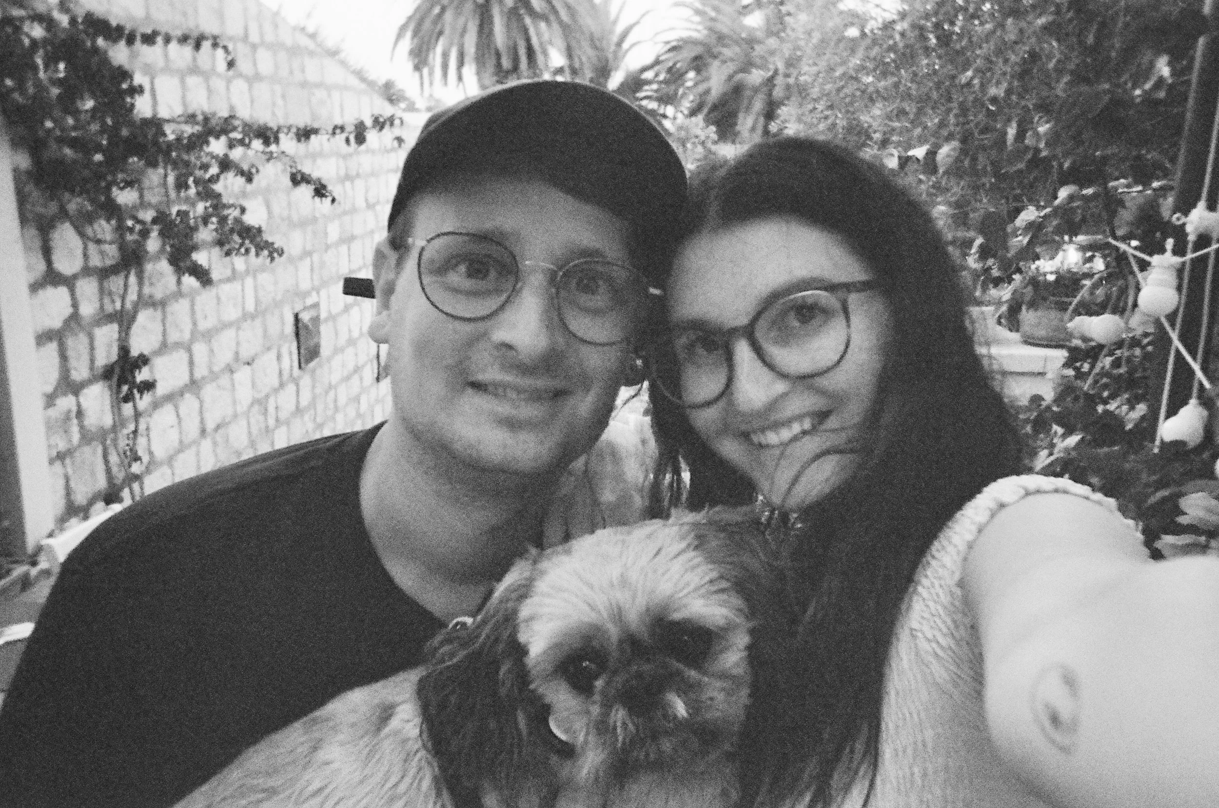 Ein schwarz-weiß Foto von einem lachenden Paar mit einem Hund zwischen ihnen, im Freien mit Pflanzen und einer Ziegelmauer im Hintergrund.