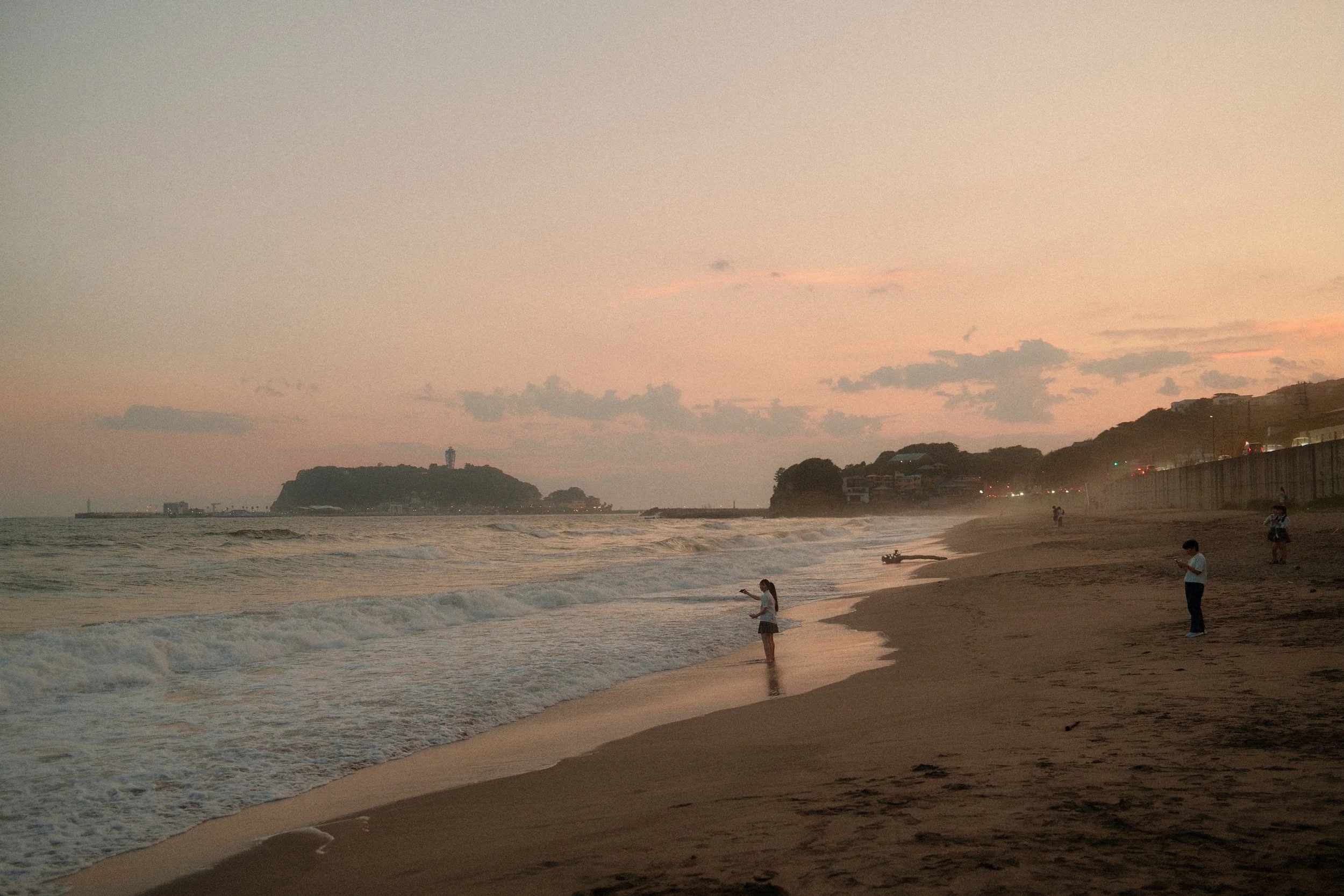 Strand bei Sonnenuntergang mit Menschen, die am Wasser spielen und fotografieren, im Hintergrund Hügel und Häuser, leicht bewölkter Himmel.