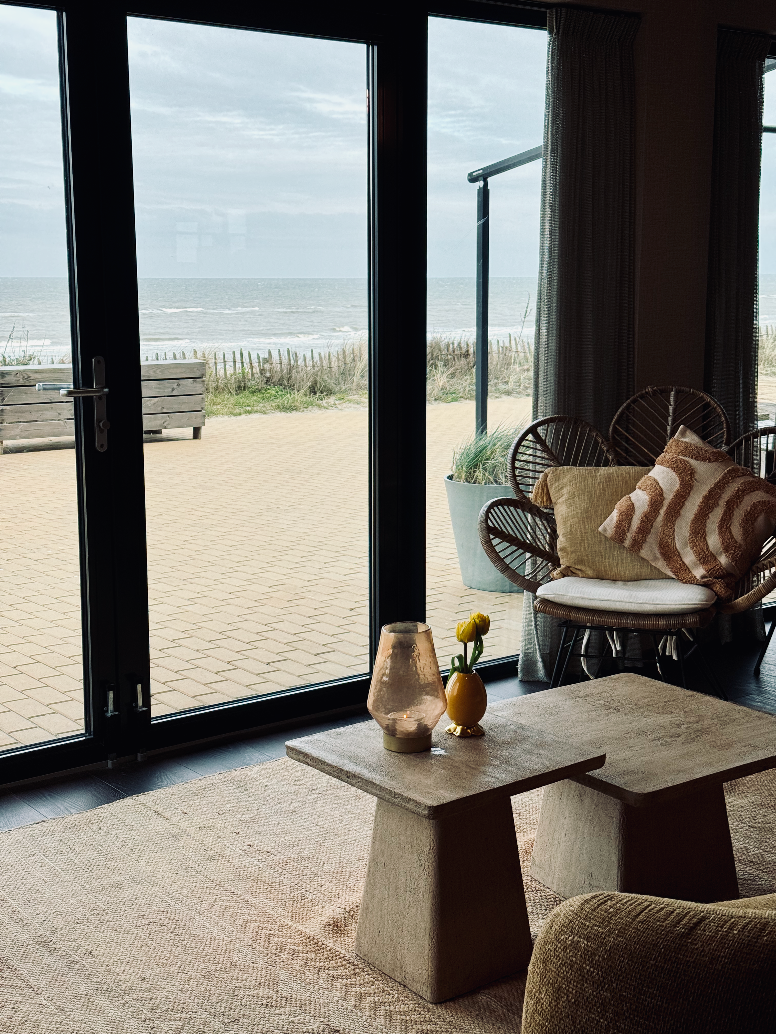 innenraum eines strandhotels mit sessel, couchtisch und großen glastüren mit direktem blick auf dünen, strand und nordsee.