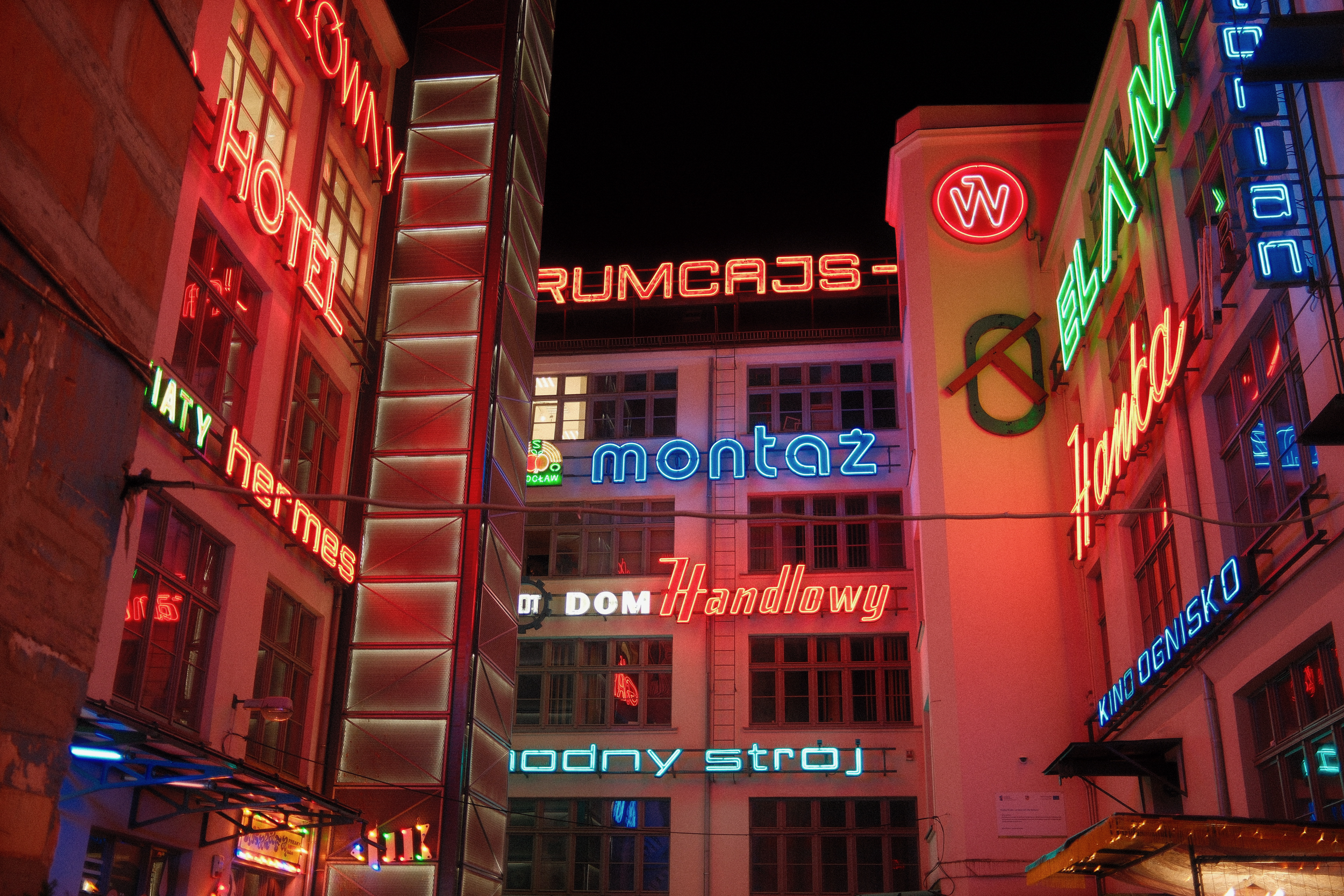 nächtlicher innenhof mit vielen bunten neonreklamen an den hausfassaden; sichtbar sind unter anderem die schriftzüge „montaż“, „dom handlowy“, „modny strój“ und „kino“, alles in rot, blau und grün vor schwarzem himmel.