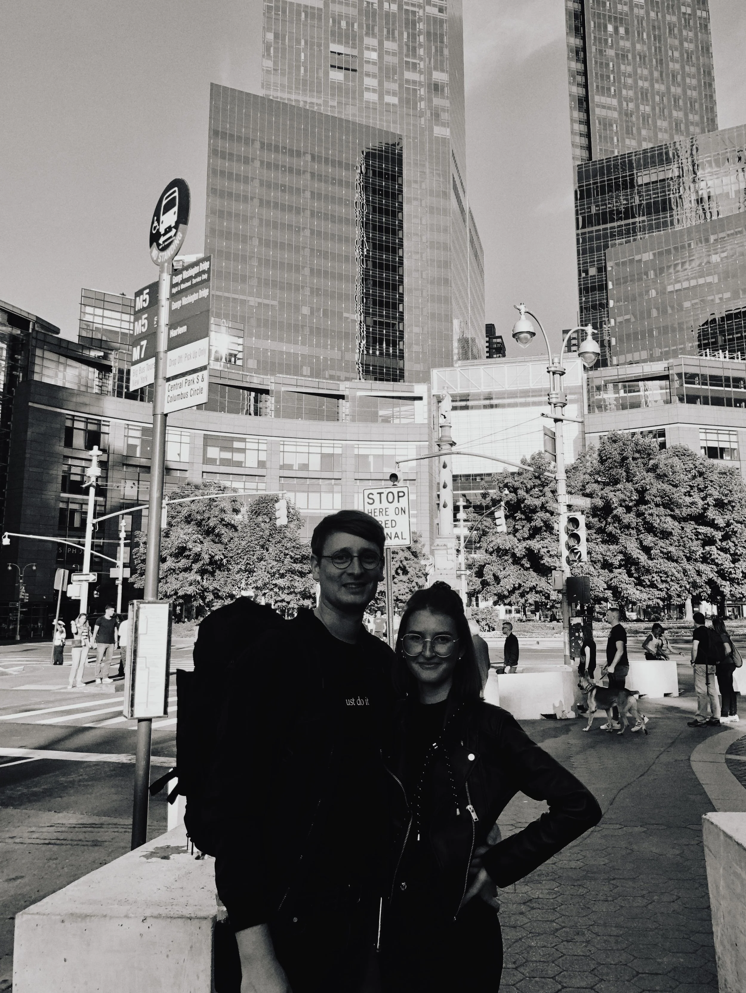 Zwei junge Menschen stehen auf einem Stadtplatz, im Hintergrund moderne Bürogebäude und Bäume, einige Passanten mit Hunden gehen vorbei.