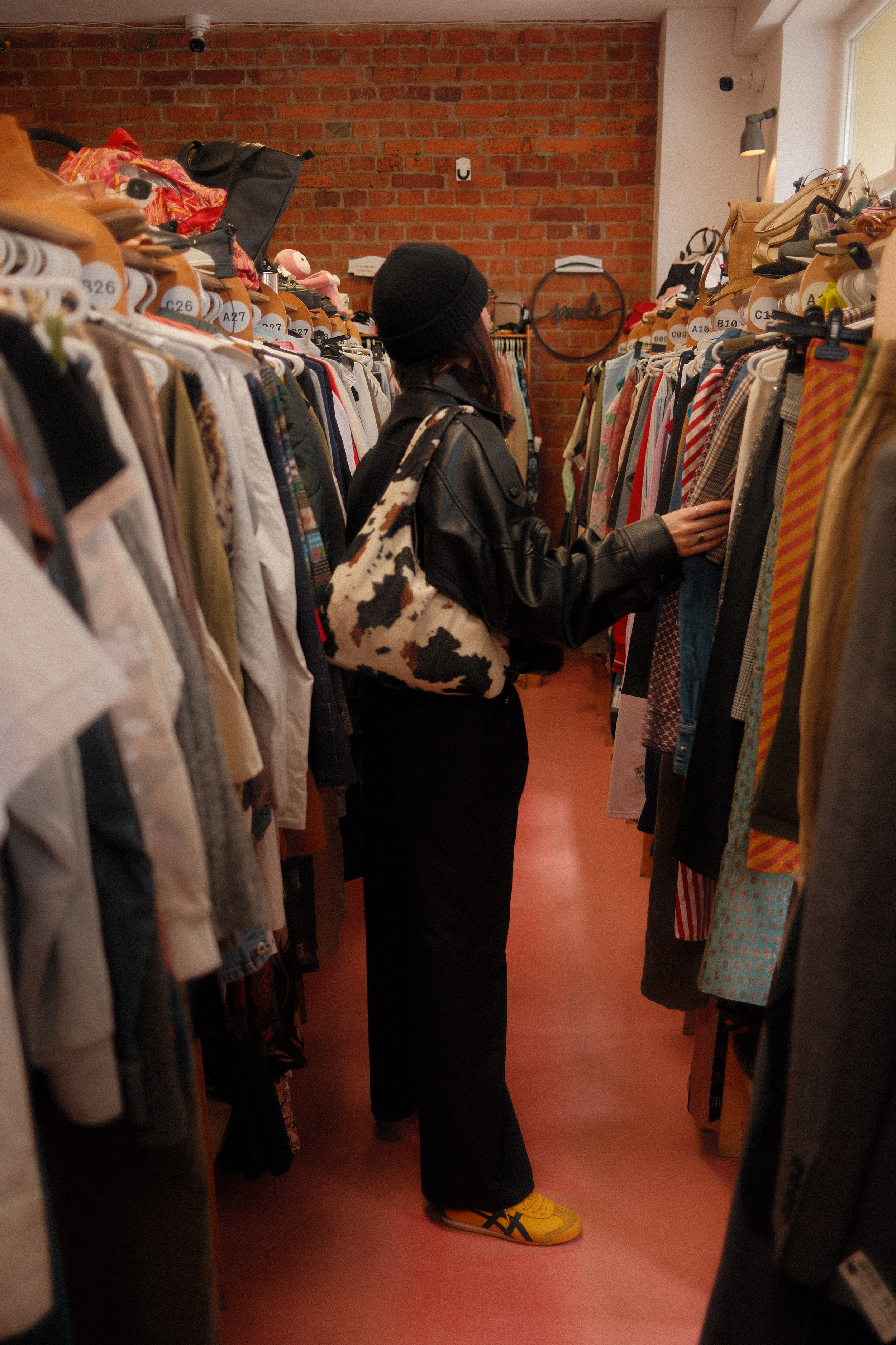 person mit schwarzer mütze und lederjacke steht zwischen zwei kleiderstangen in einem secondhand-laden, greift nach einem kleidungsstück; roter boden, backsteinwand im hintergrund, oben liegen taschen und accessoires.