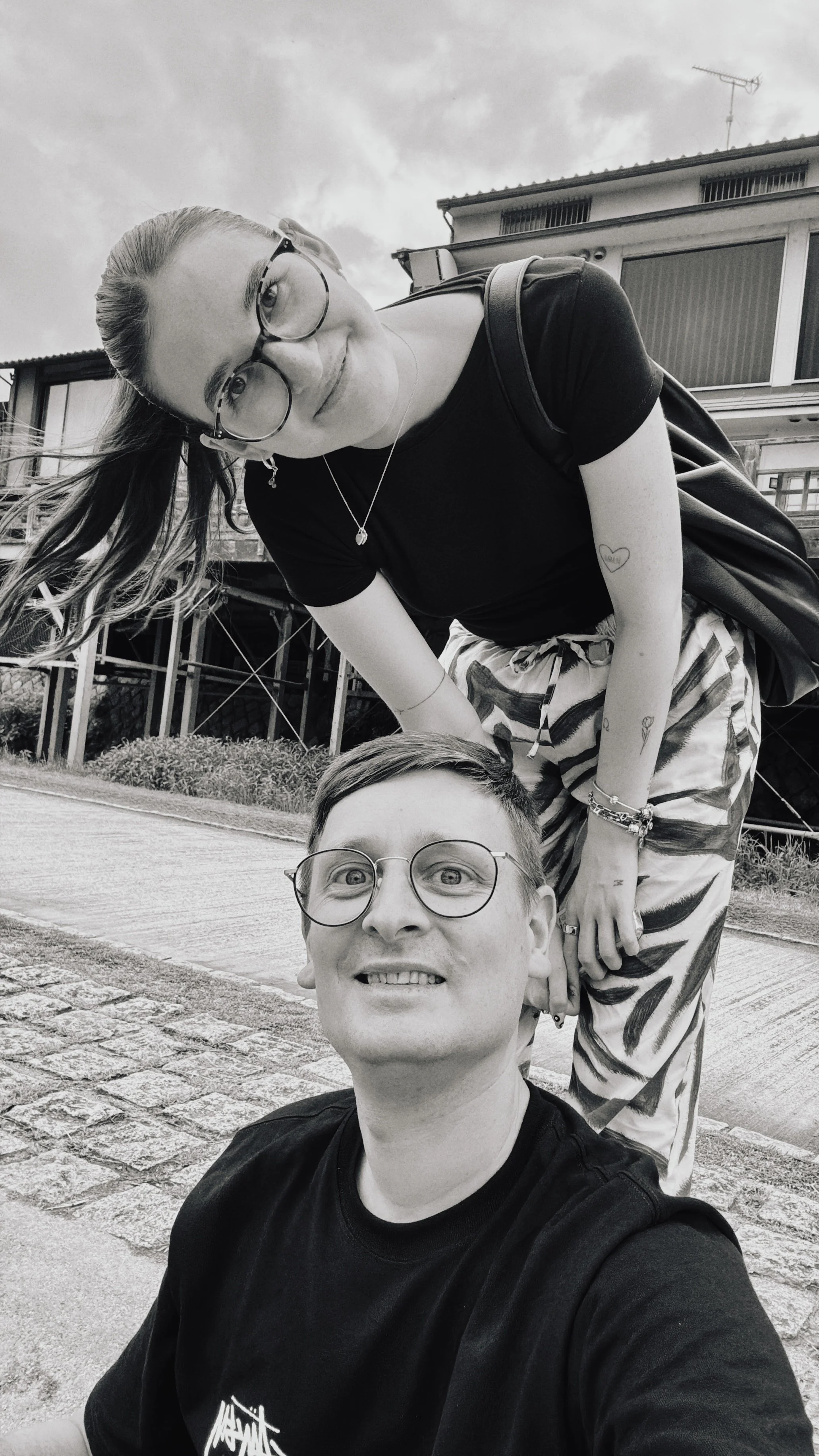 Ein Schwarz-Weiß-Foto von einem Paar, das auf einer Straße im Freien Fotos macht. Die Frau trägt eine schwarze T-Shirt, eine Brille, mehrere Armbänder, Tattoos und trägt eine Tasche. Der Mann trägt eine schwarze T-Shirt, eine Brille, und hat kurze Haare. Im Hintergrund sind Häuser und ein bewölkter Himmel zu sehen.