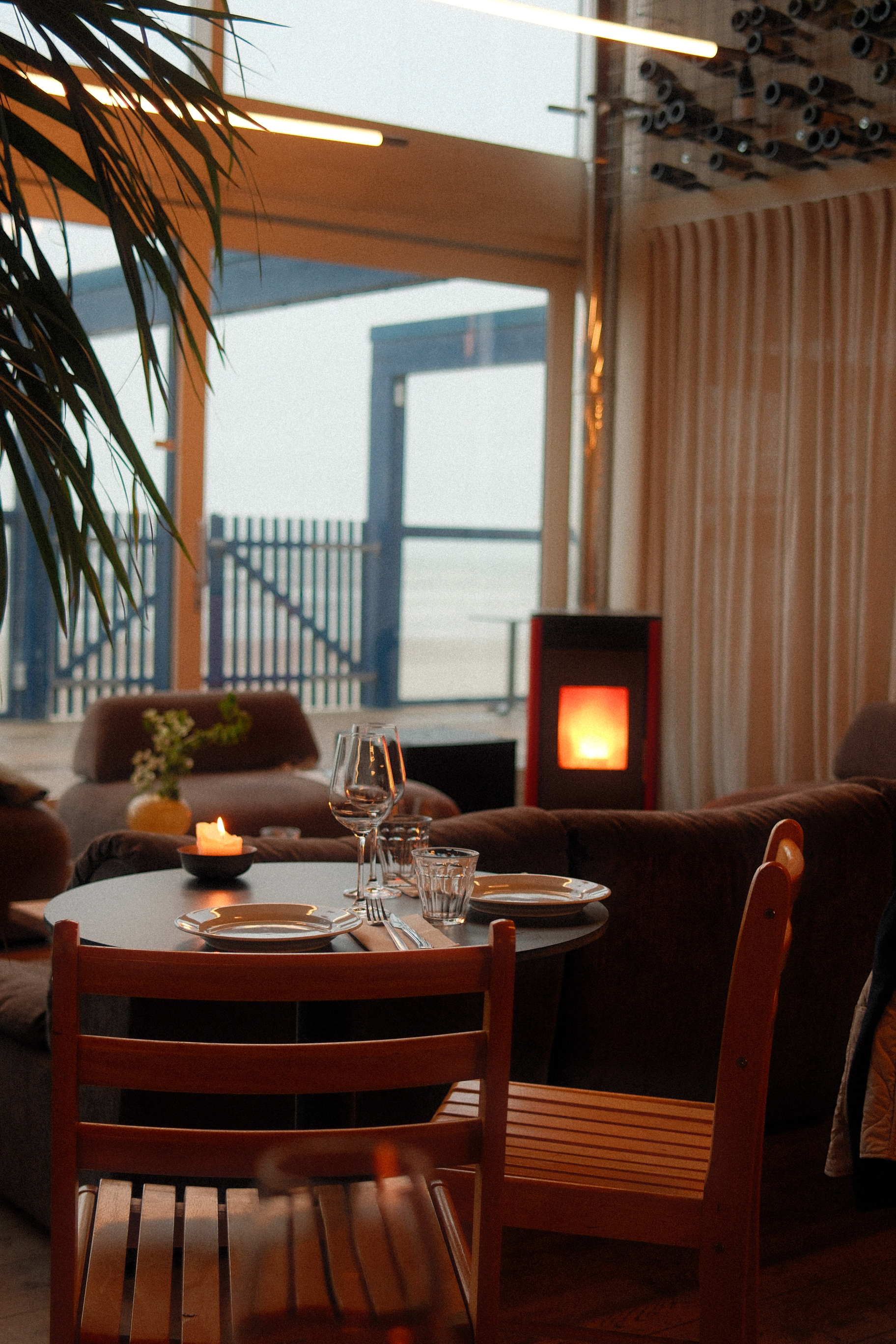 innenraum eines strandrestaurants in zandvoort mit gedecktem tisch, warmem kaminlicht und blick durch große fenster Richtung meer.
