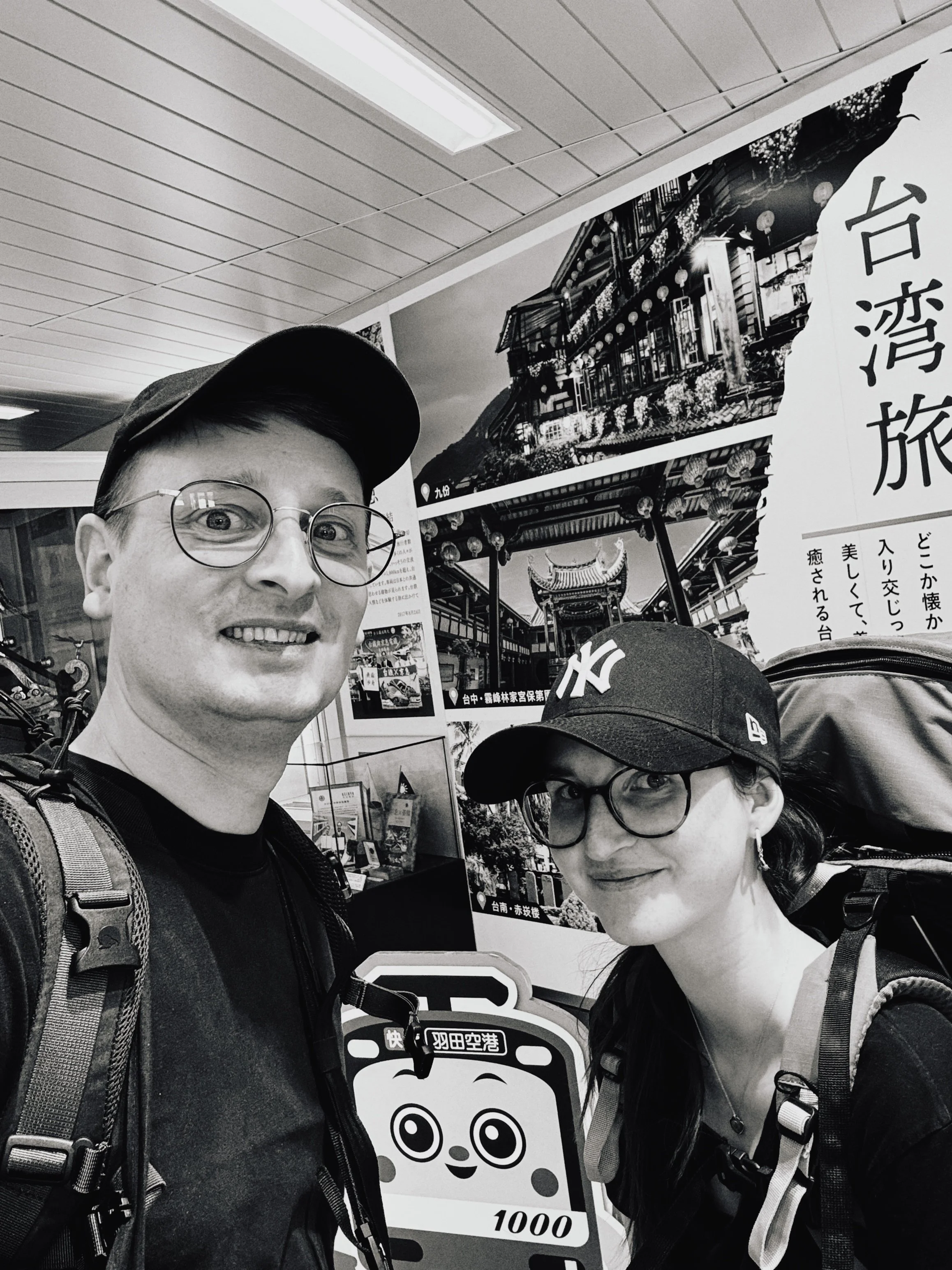 Zwei reisende Personen, ein Mann und eine Frau, posieren mit Rucksäcken vor einem Poster mit chinesischer Schrift und Bildern von traditionellen Gebäuden in Taiwan.