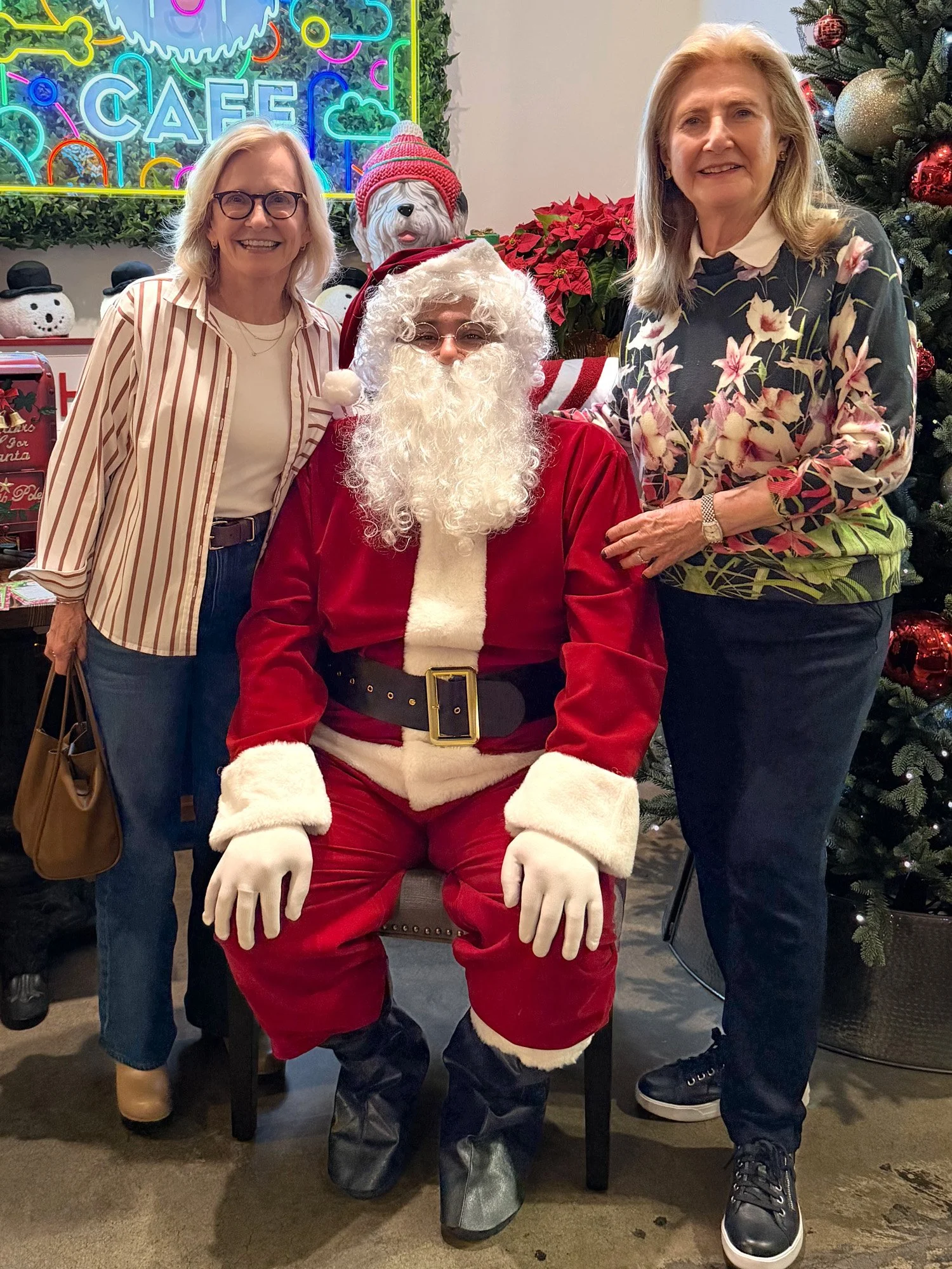 Two women and Santa Claus sitting between them pose for a photo in a festive holiday setting with Christmas decorations, including a Christmas tree, poinsettias, and holiday-themed figures.