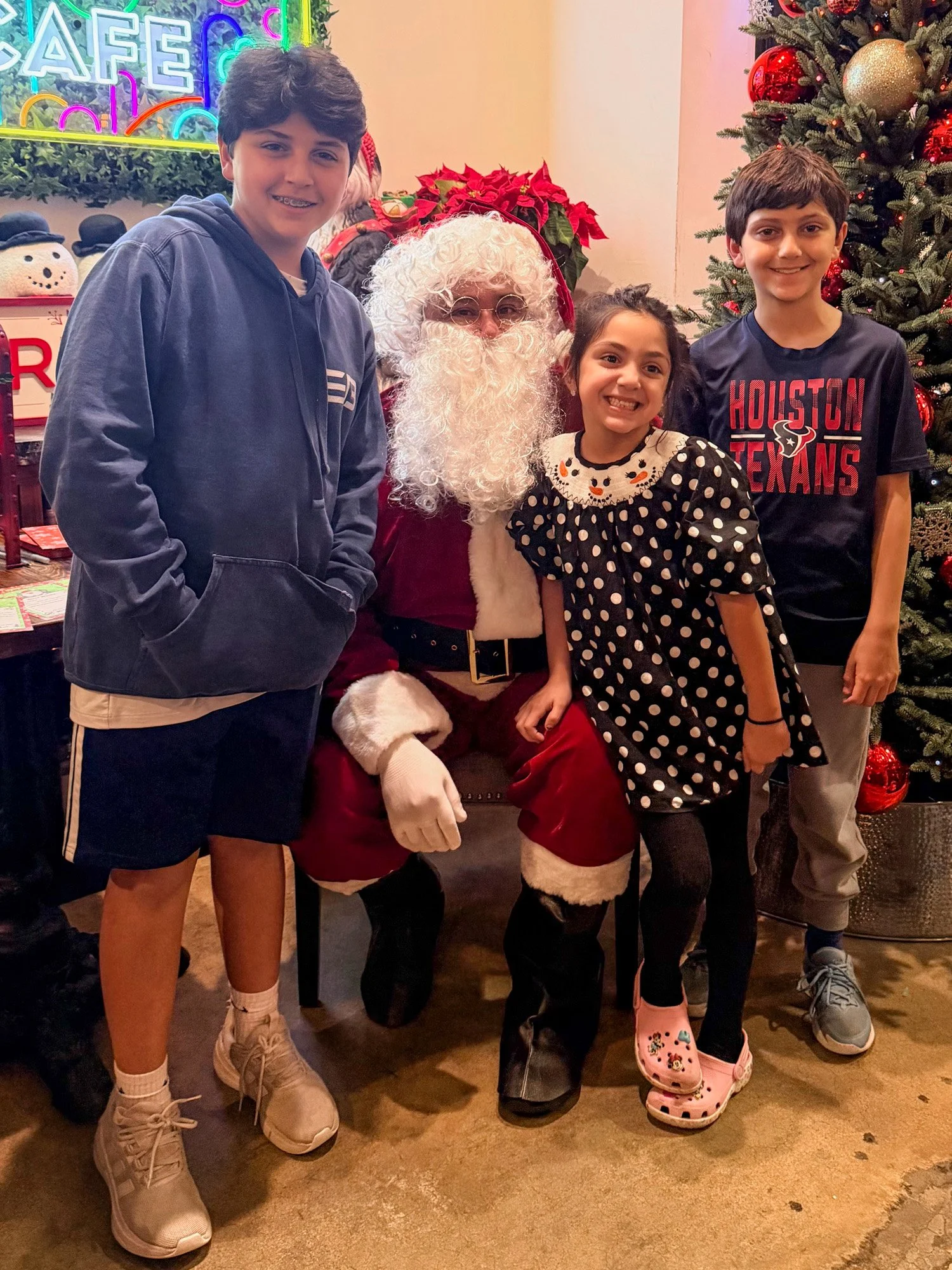 Three children posing with Santa Claus next to a decorated Christmas tree and poinsettia plant, in front of a neon sign that reads 'Cafe'.