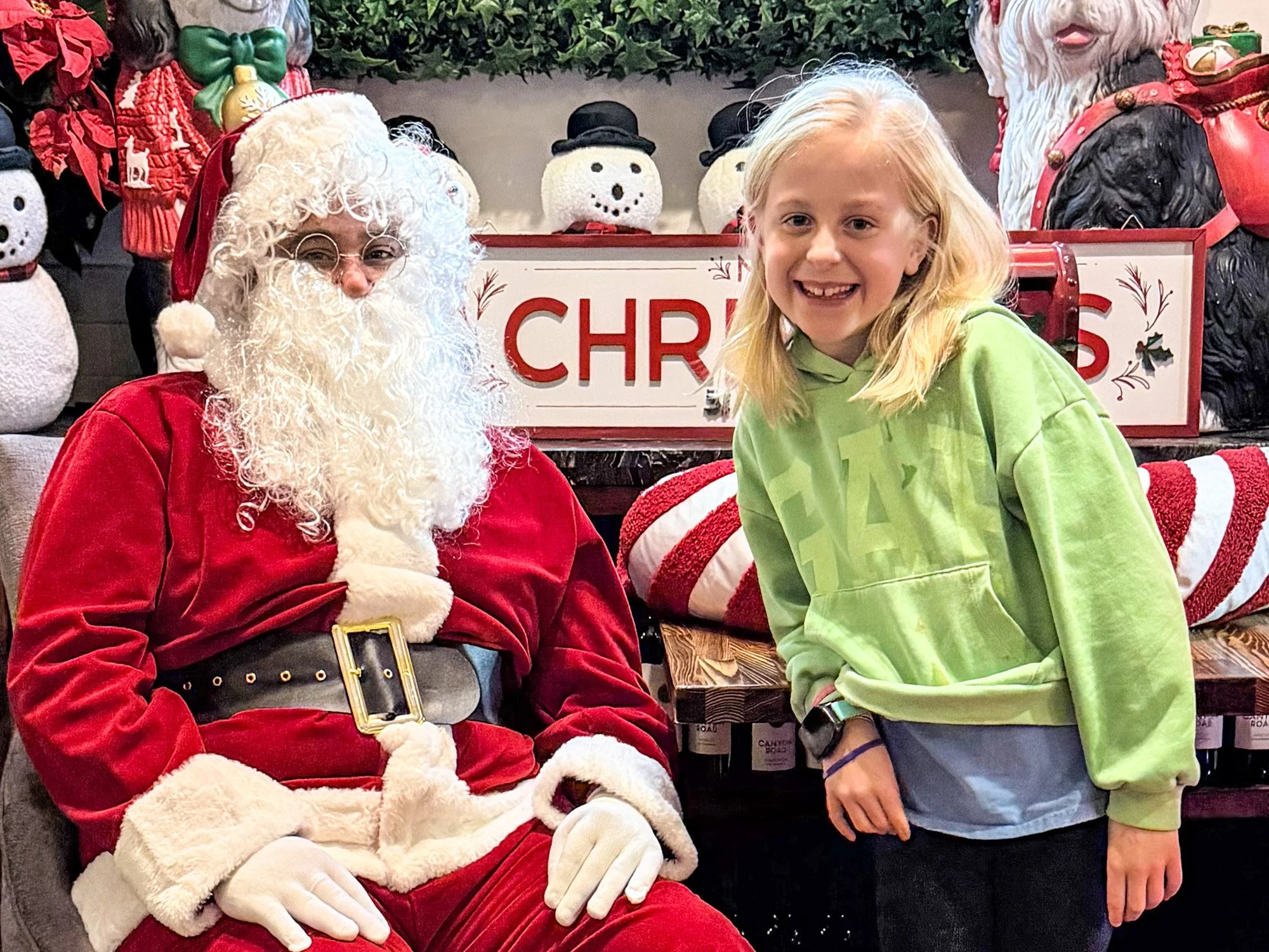 A young girl with blonde hair smiling and standing next to a person dressed as Santa Claus, surrounded by Christmas decorations including snowman figures and a sign that says 'Christmas'.