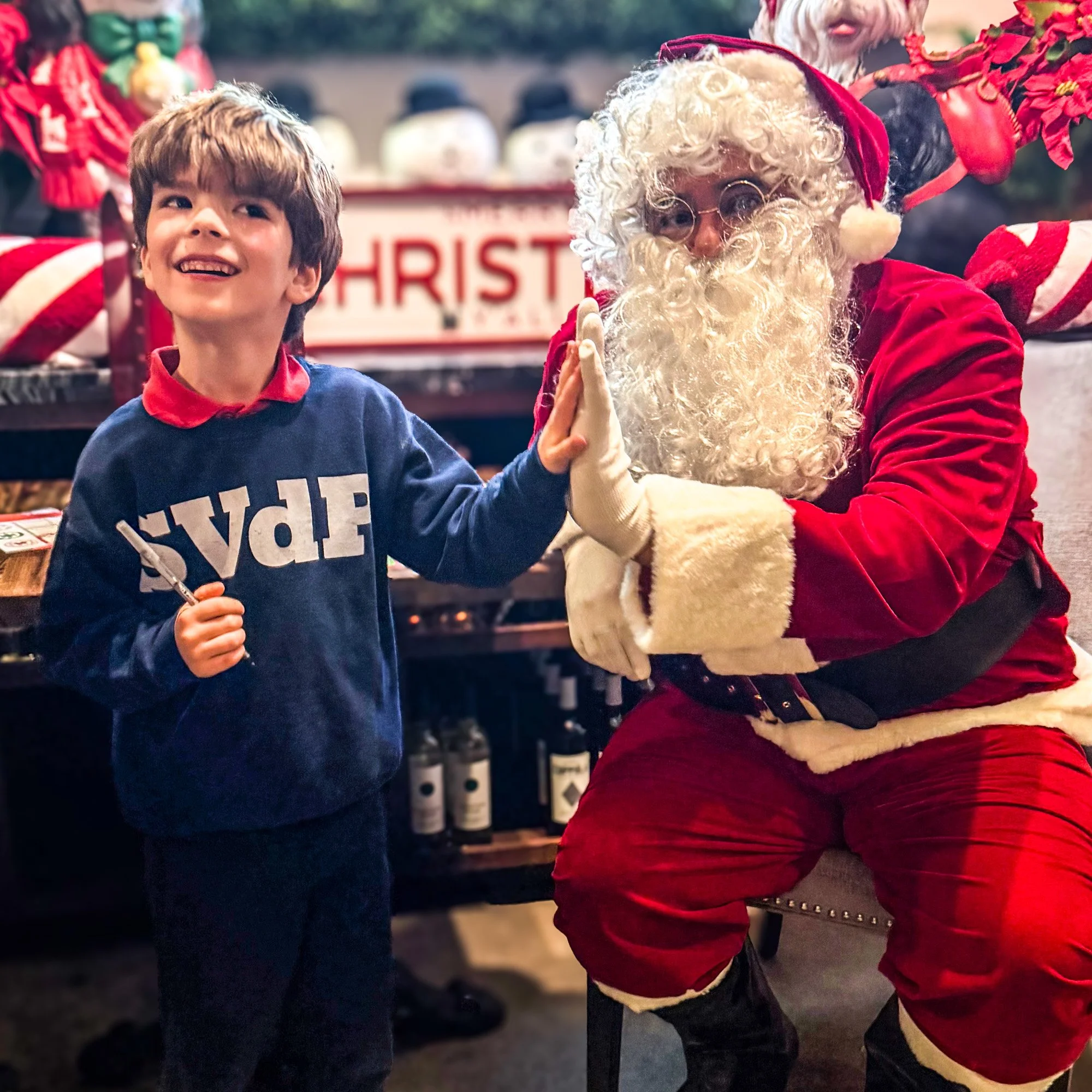 A young boy in a blue sweater giving a high five to Santa Claus, who is dressed in a red suit with white fur trim and glasses. They are smiling, and in the background, Christmas decorations and a sign that says 'CHRISTMAS' are visible.