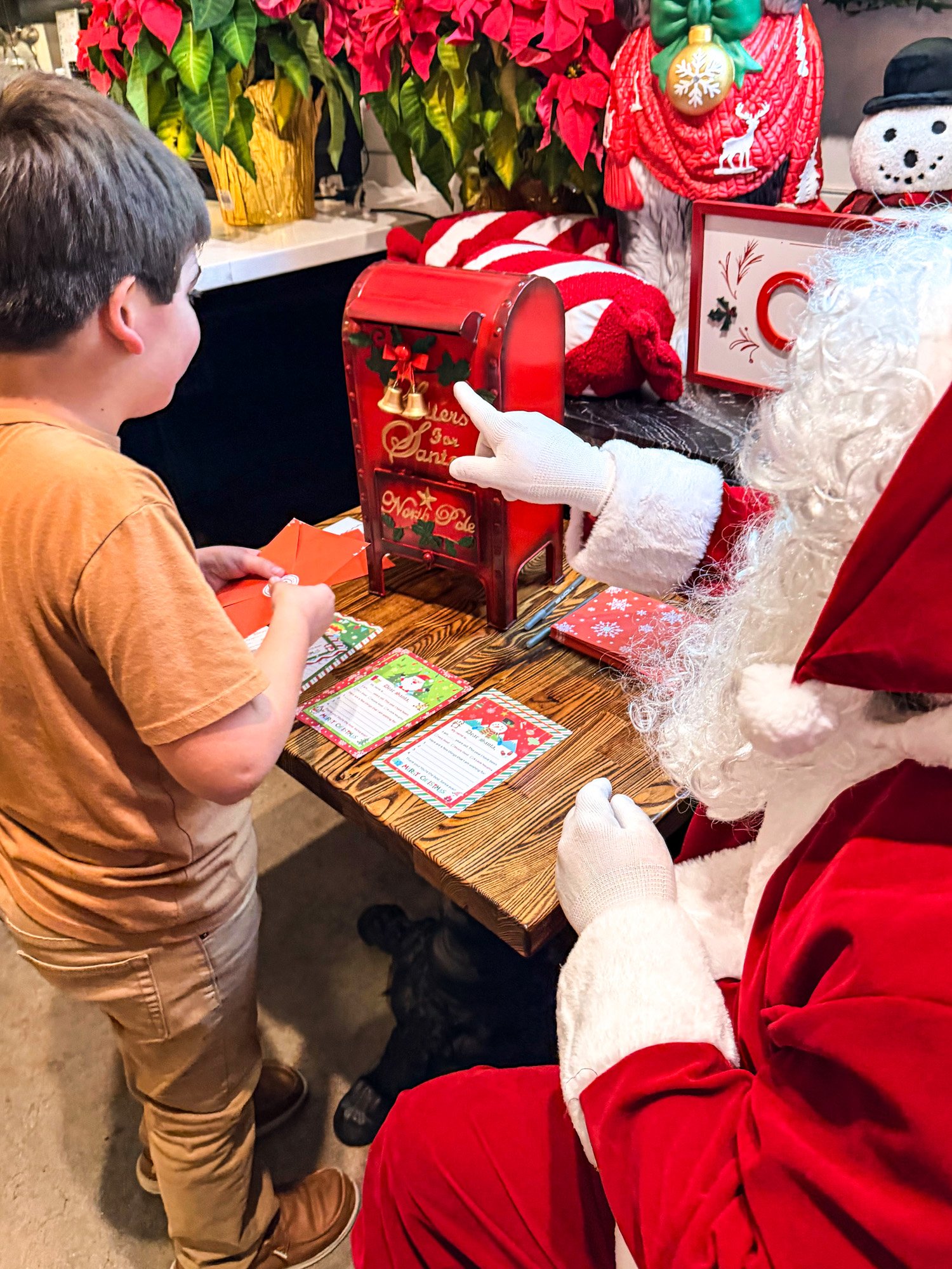 A child receiving a Christmas card from Santa Claus at a holiday event, with Christmas decorations and a mailbox on the table.