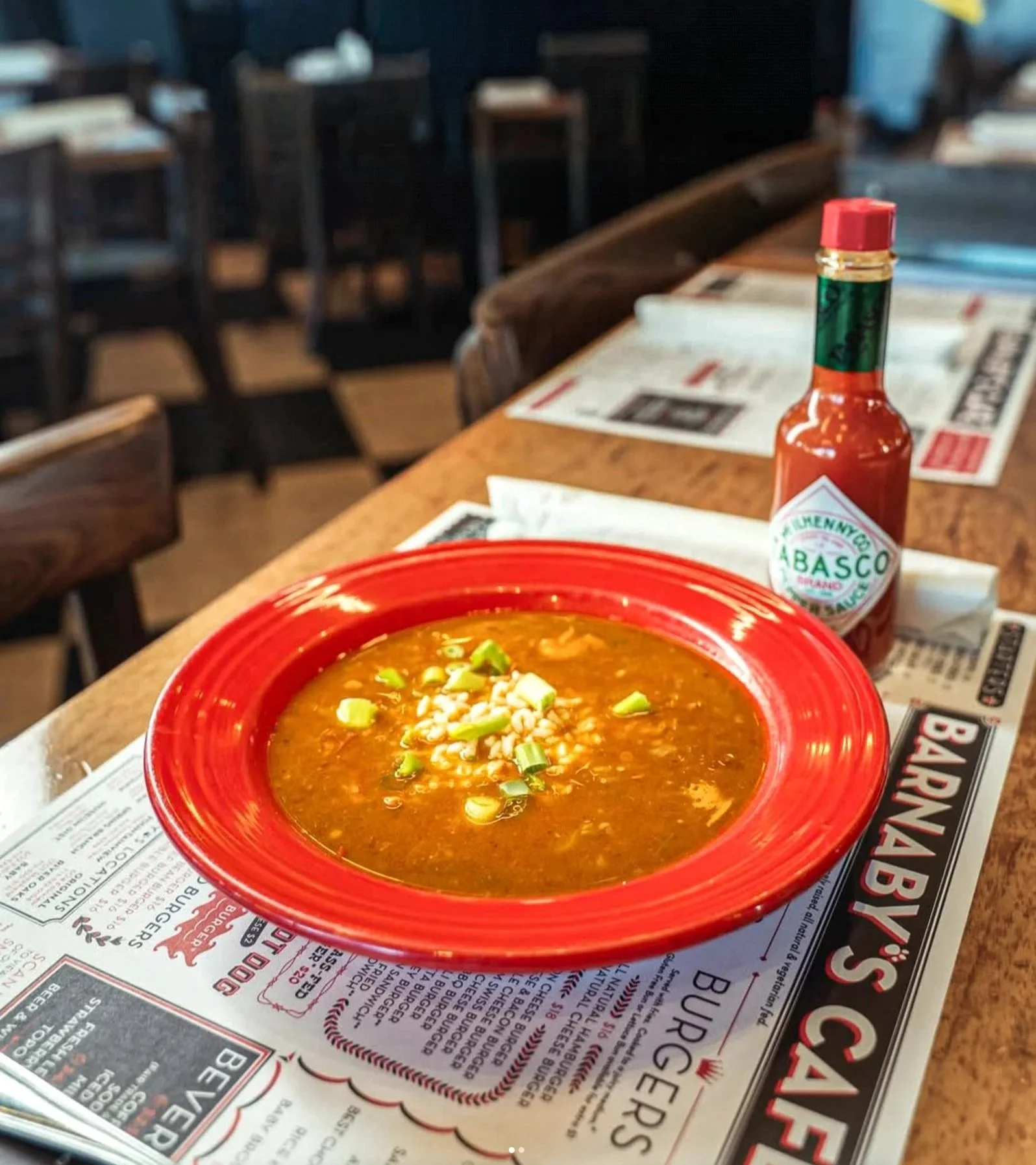 A bowl of chili topped with chopped green onions on a red plate, with a bottle of Tabasco sauce on the table in a restaurant setting.