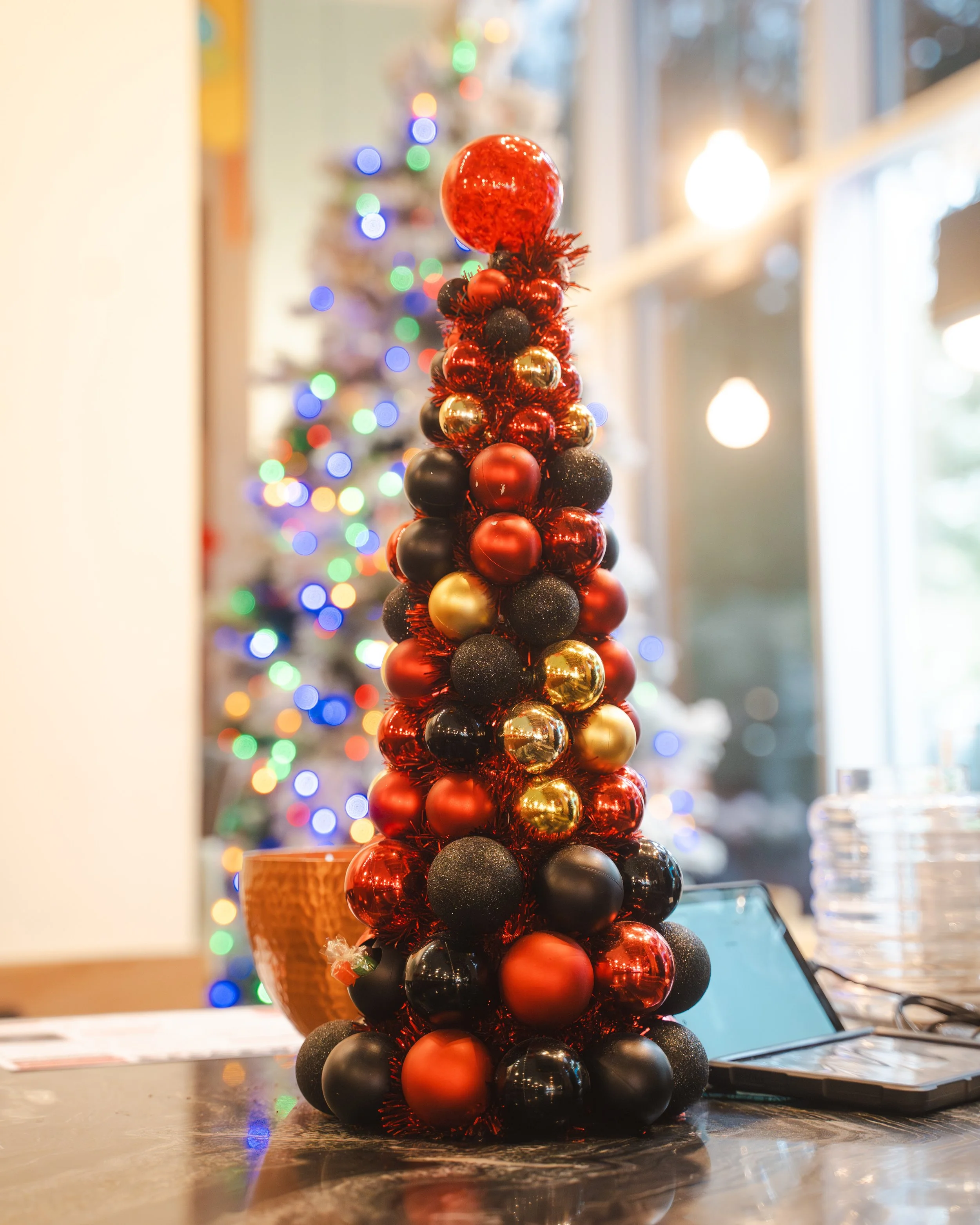 A small Christmas tree decoration made of red, black, and gold Christmas ornaments stacked on a table, with a larger decorated Christmas tree with colorful lights in the blurred background.
