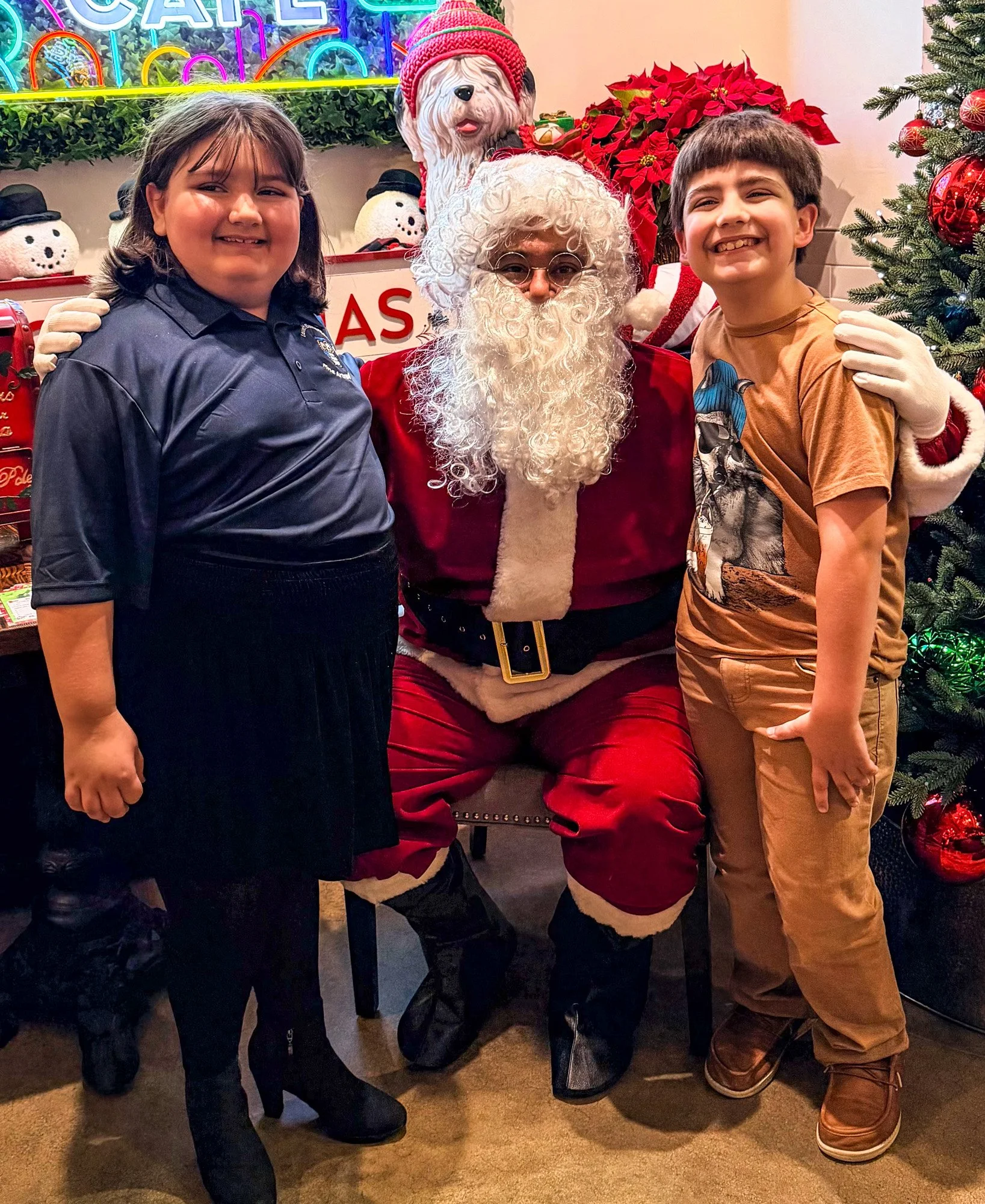 Two children smiling and posing with Santa Claus at a Christmas event. The girl on the left has shoulder-length dark hair and is wearing a navy shirt and black skirt. The boy on the right has short dark hair, is wearing a brown T-shirt with a graphic