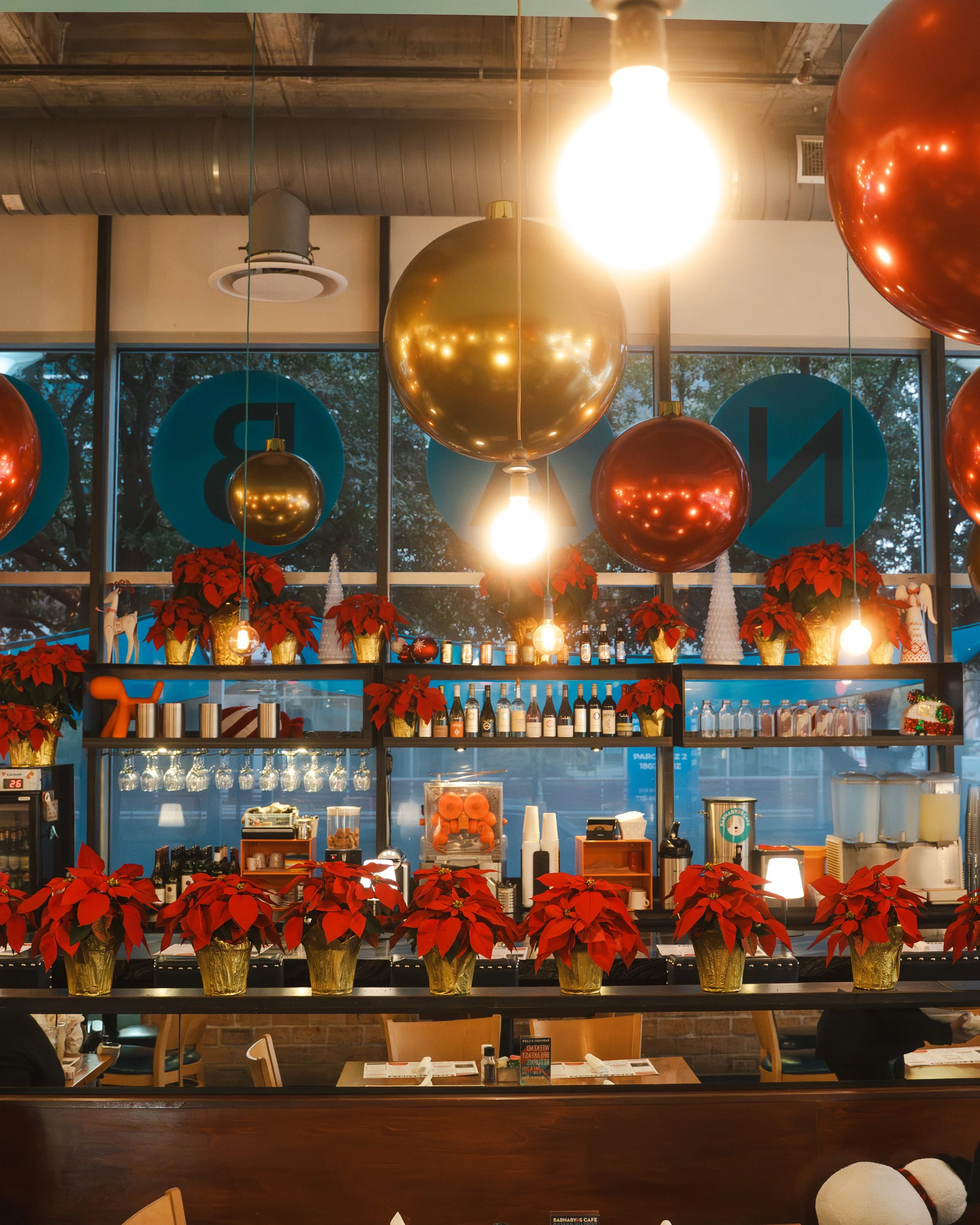 Decorated cafe with hanging large and small gold and red balloons, red poinsettias in gold pots, and a bar area with bottles, glasses, and holiday themed decor.