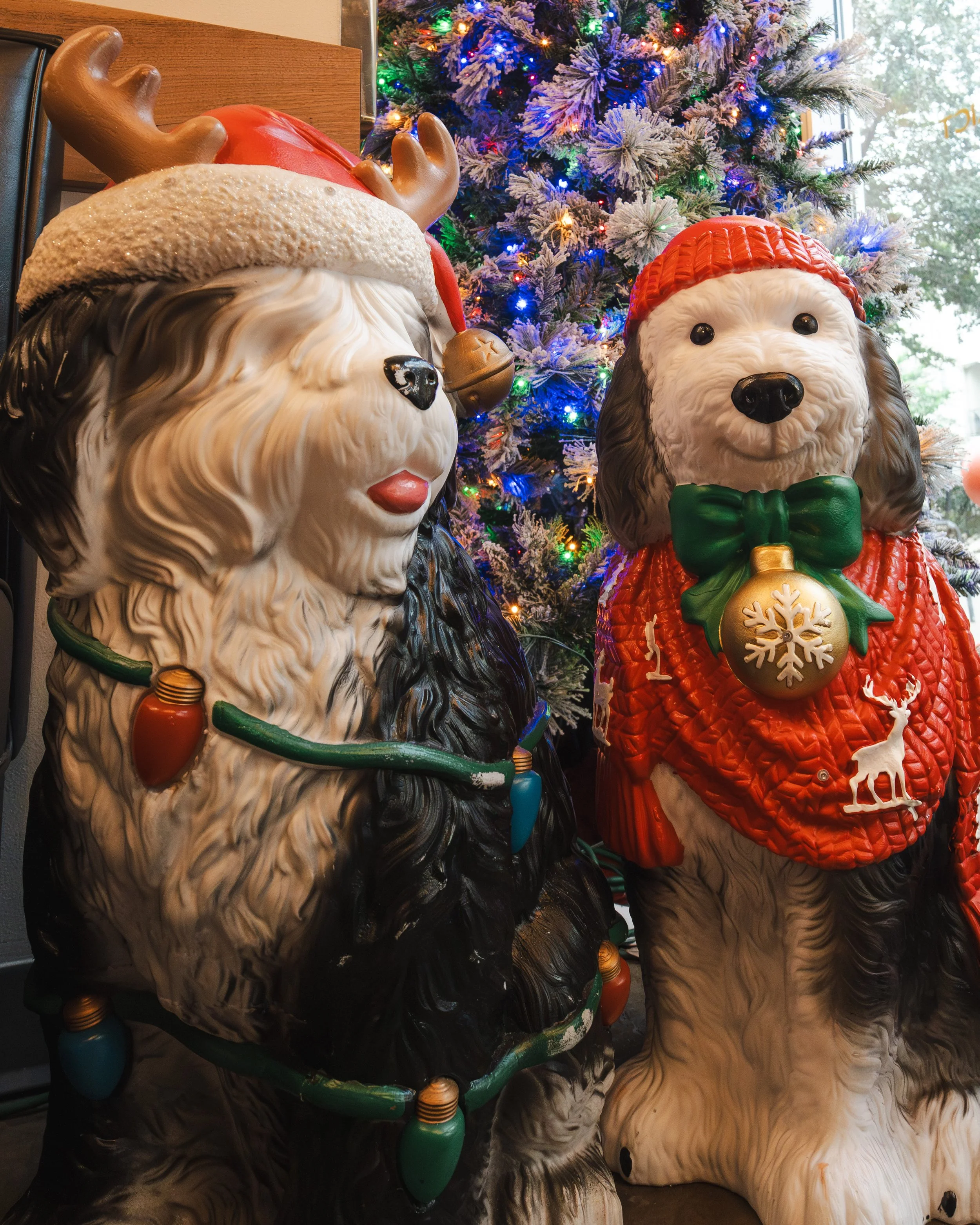 Decorative Christmas statues of two dogs, one with a reindeer antler hat and the other wearing a red sweater and a green bow tie, in front of a decorated Christmas tree with colorful lights.
