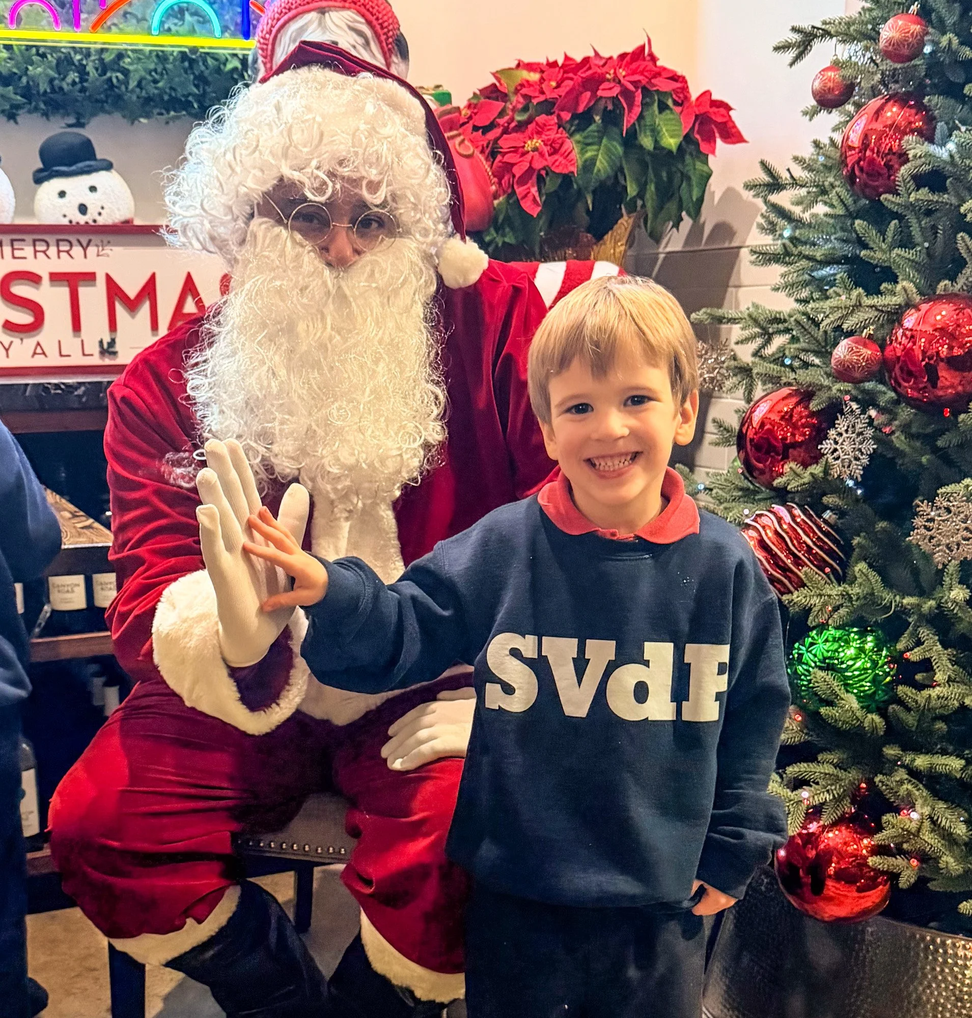 A smiling young boy giving a high five to Santa Claus during a Christmas event, standing next to a decorated Christmas tree with red ornaments and lights.