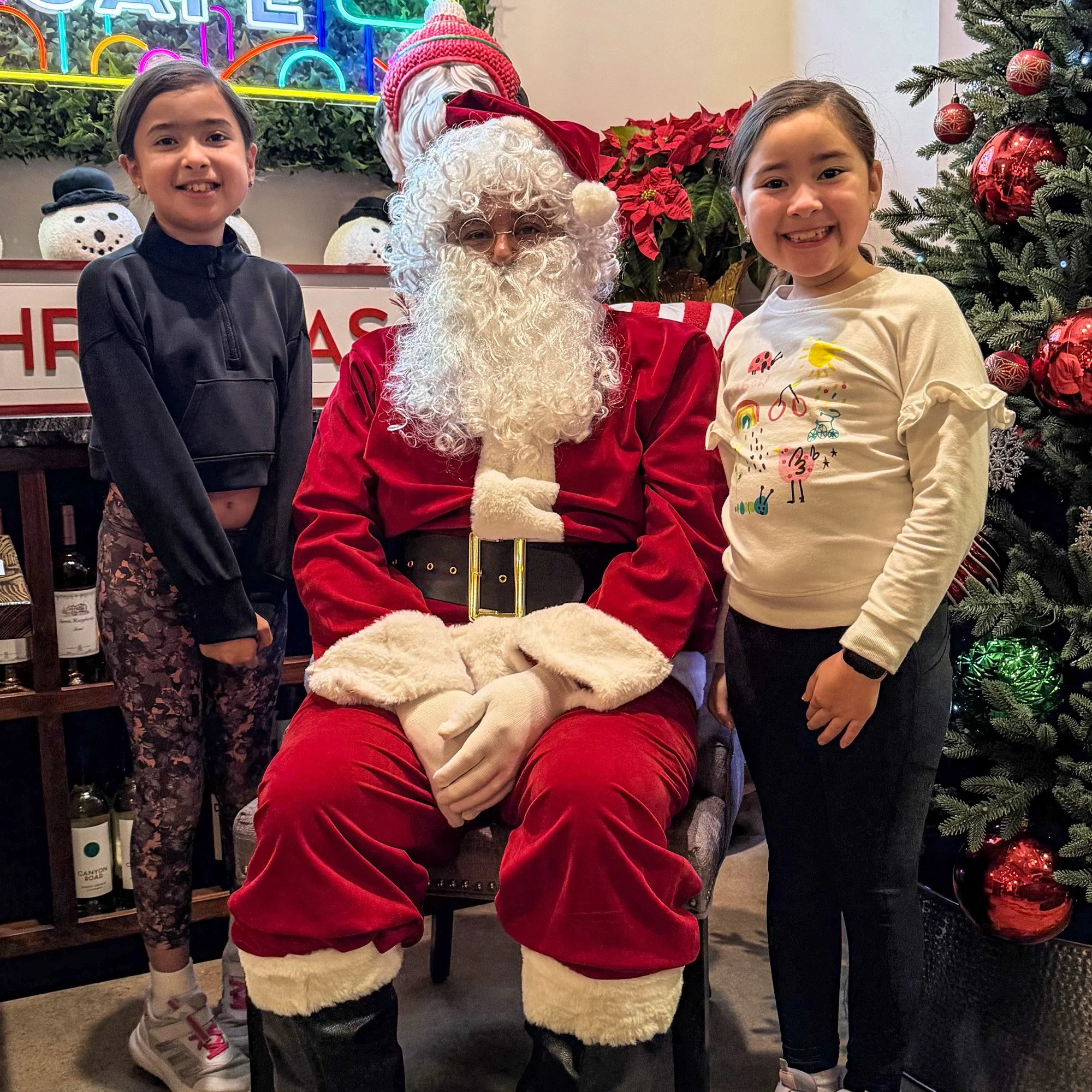 Two young girls standing beside Santa Claus, who is seated. They are posing for a photo in a festive setting with Christmas decorations, including a decorated Christmas tree and snowman ornaments.