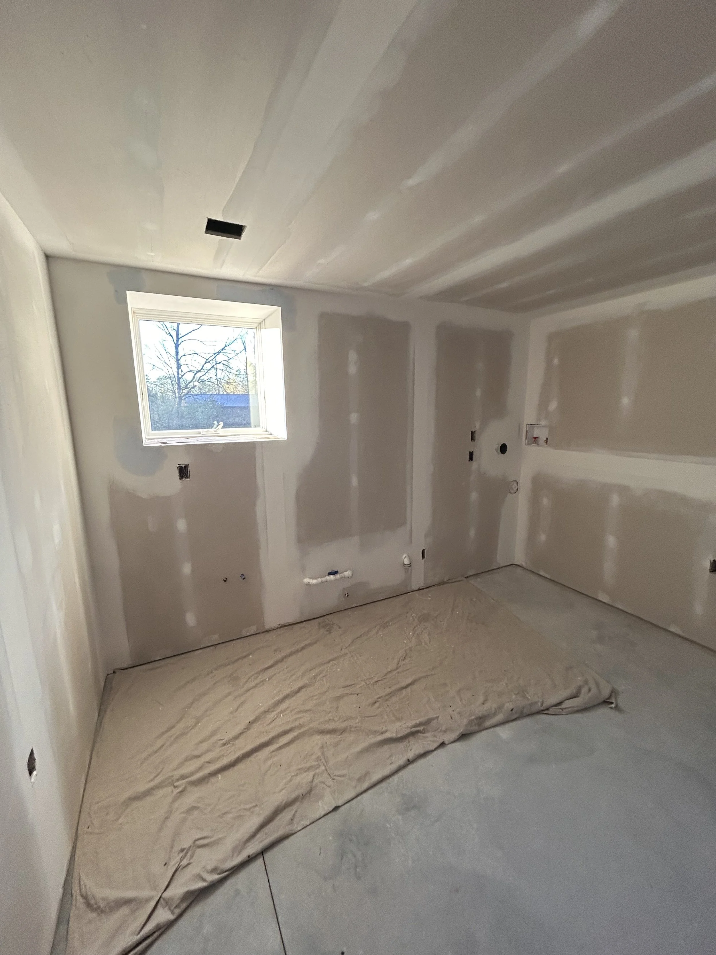 An unfinished room under construction with drywall installed on walls and ceiling, a small window, and a sheet of drywall on the floor.