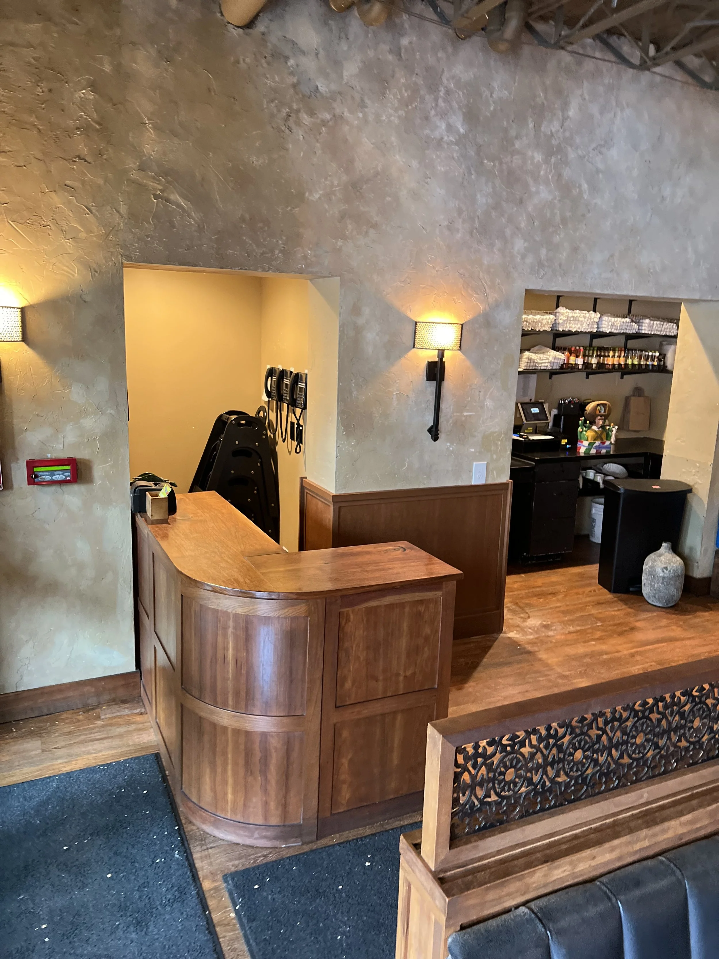 Inside a restaurant, view of a wooden bar counter, a coffee station with cups and condiments, and a customer service area with a computer and shelves of supplies, decorated with warm yellow wall lights.