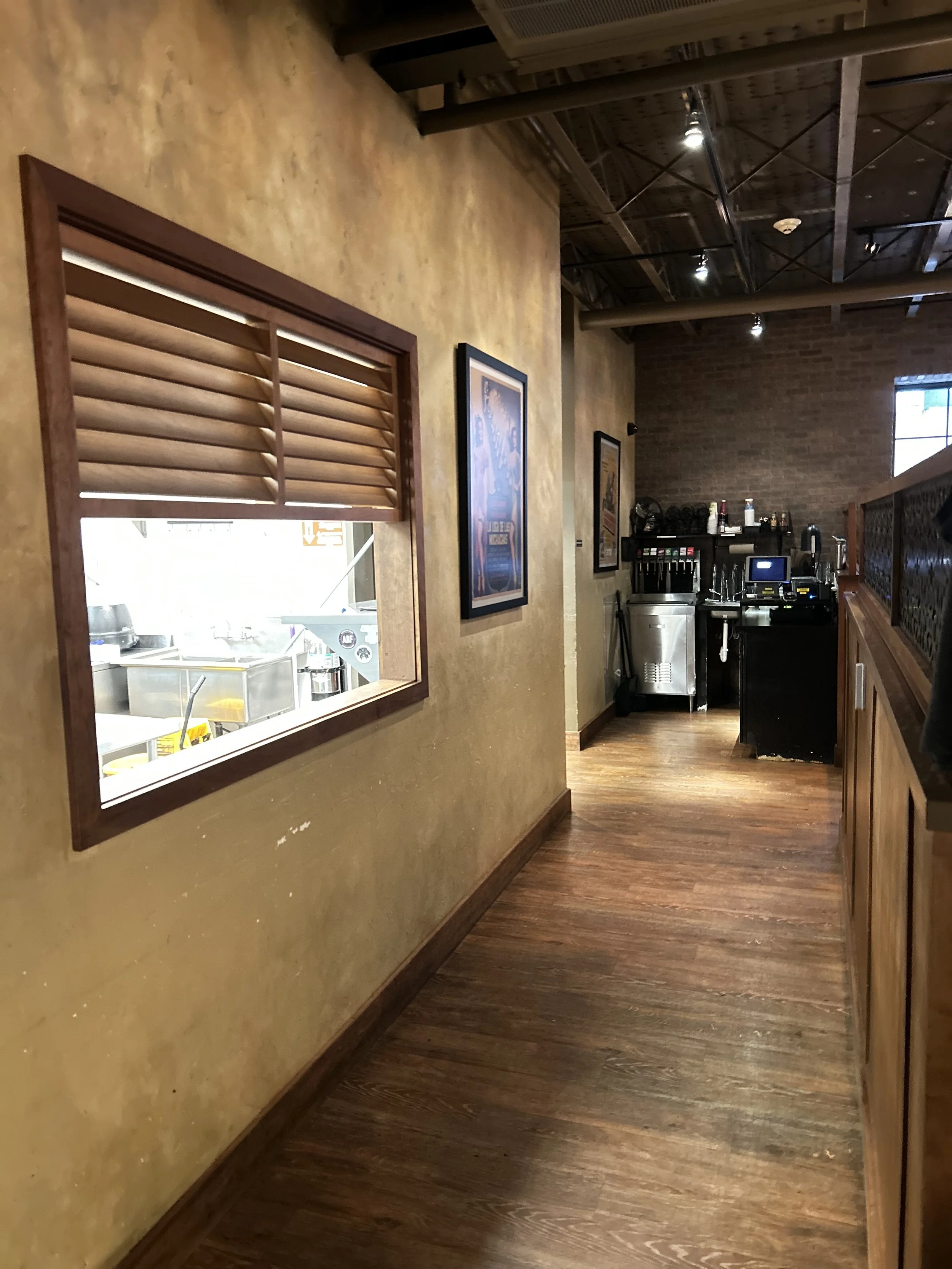 Interior view of a restaurant kitchen area through a window in the wall, with a beer dispenser and kitchen equipment in the background, and framed posters on the beige painted wall.