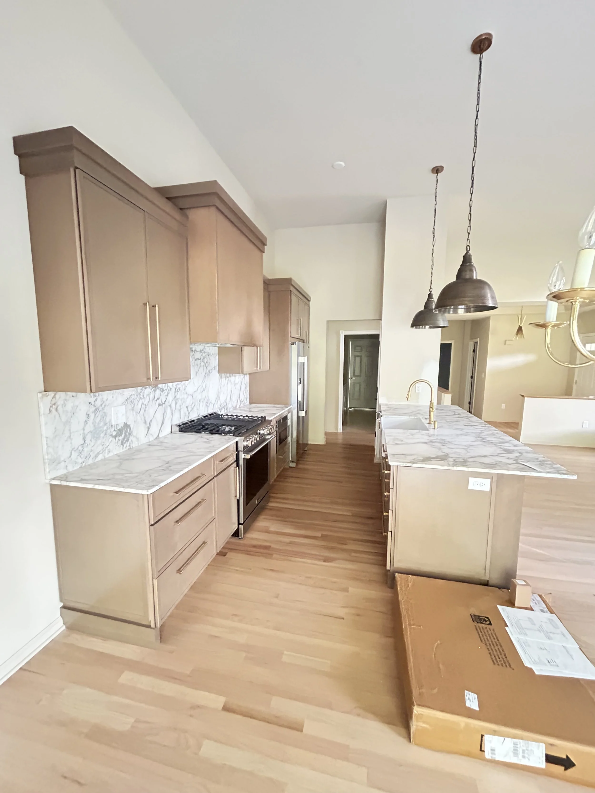 Modern kitchen with beige cabinets, marble countertops, and a wooden floor. Two hanging pendant lights over the island, with a cardboard box on the floor.