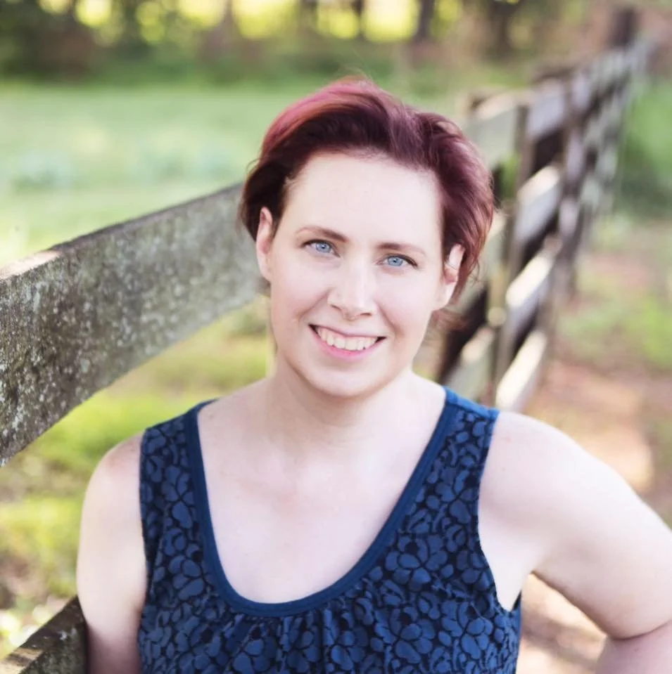 A woman with short reddish-brown hair and blue eyes smiling outdoors next to a wooden fence, wearing a dark blue sleeveless top in a natural setting.