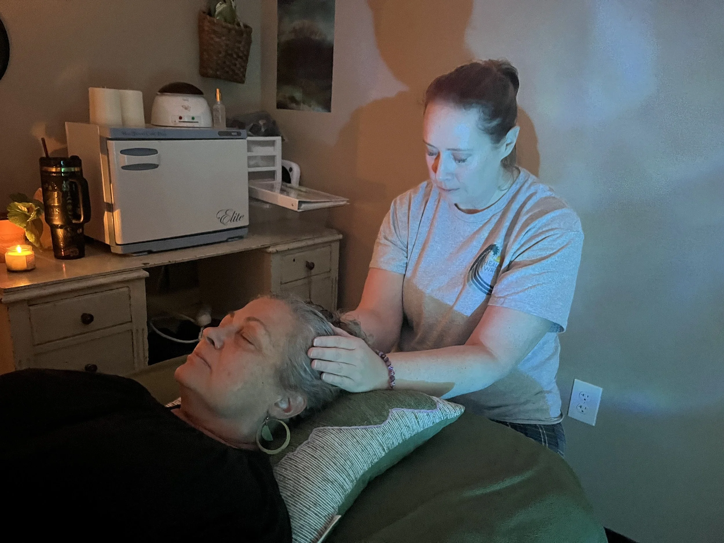 A woman giving a massage to a person lying down on a bed or massage table in a cozy room with soft lighting and candlelight.