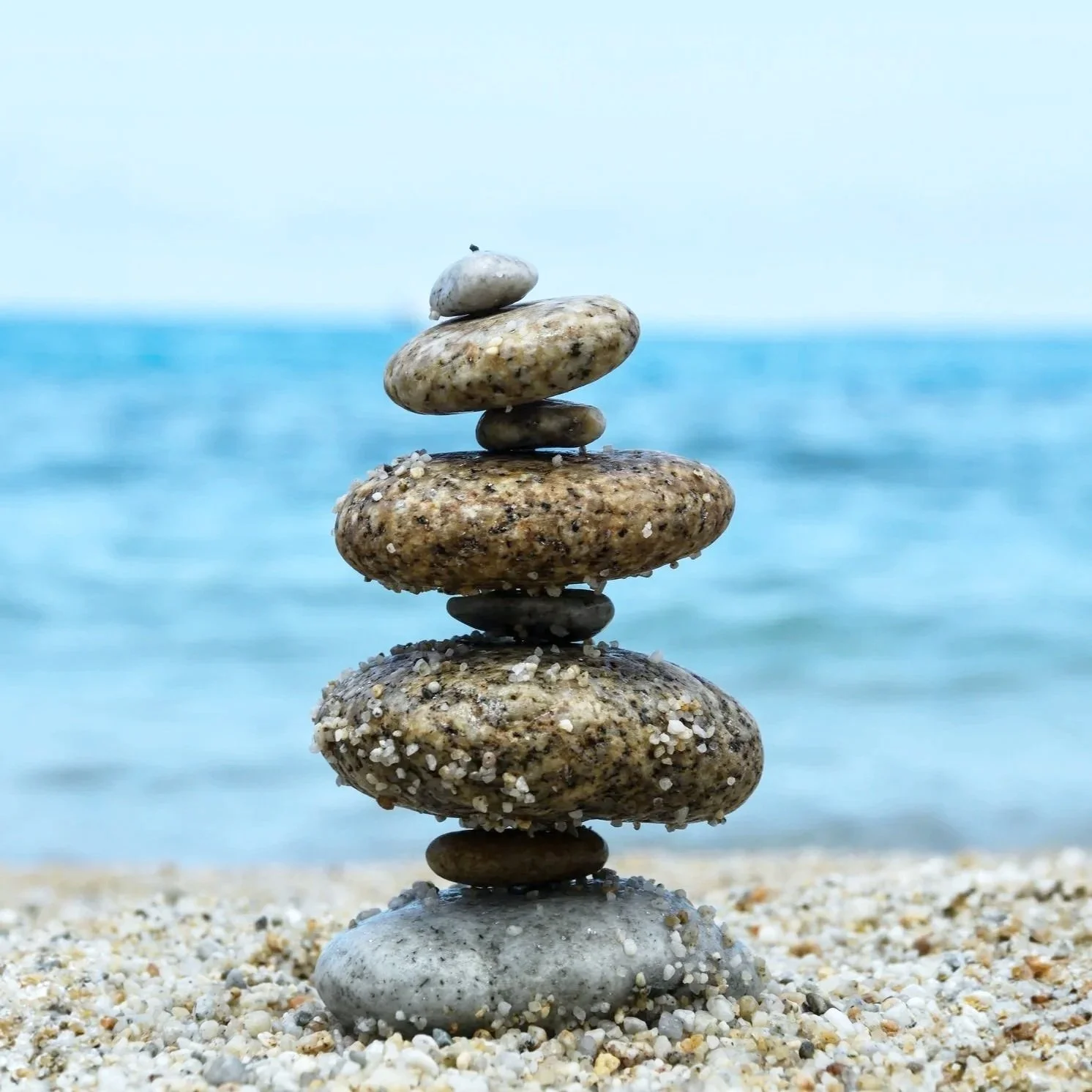 A stack of smooth, multicolored stones balanced on a sandy beach with the ocean in the background.