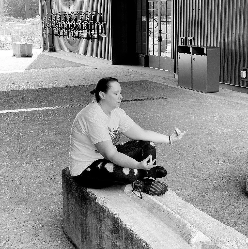 A woman practicing meditation outdoors while sitting cross-legged on a concrete block, with her eyes closed and hands in a mudra gesture.