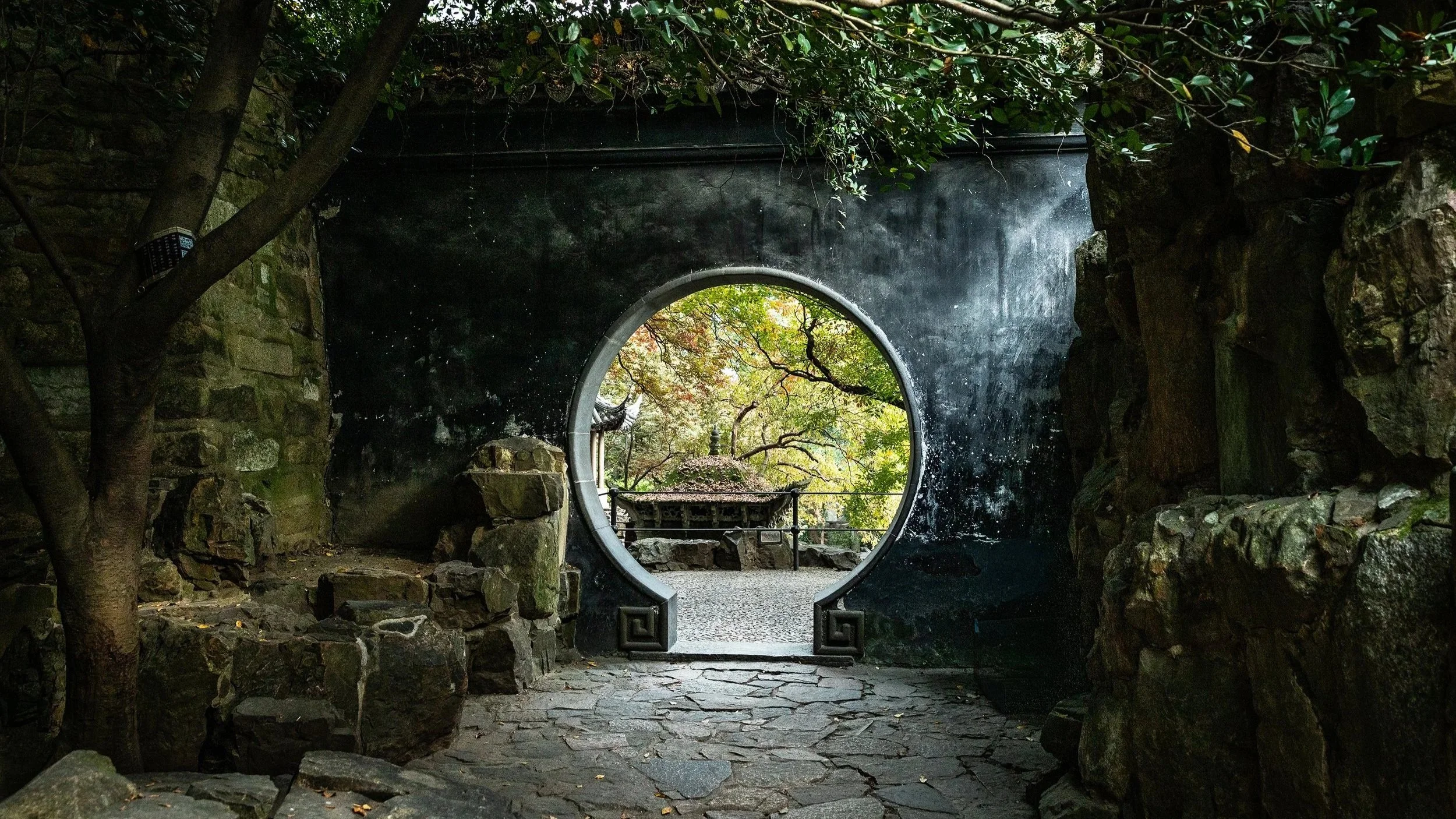 Shadowed stone archway and and circular gate opening into a secluded garden, symbolizing the threshold between worlds and inner transformation.