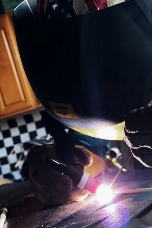 Person welding metal at a workbench indoors, wearing protective gear including a welding helmet and gloves, with a bright weld flame visible.