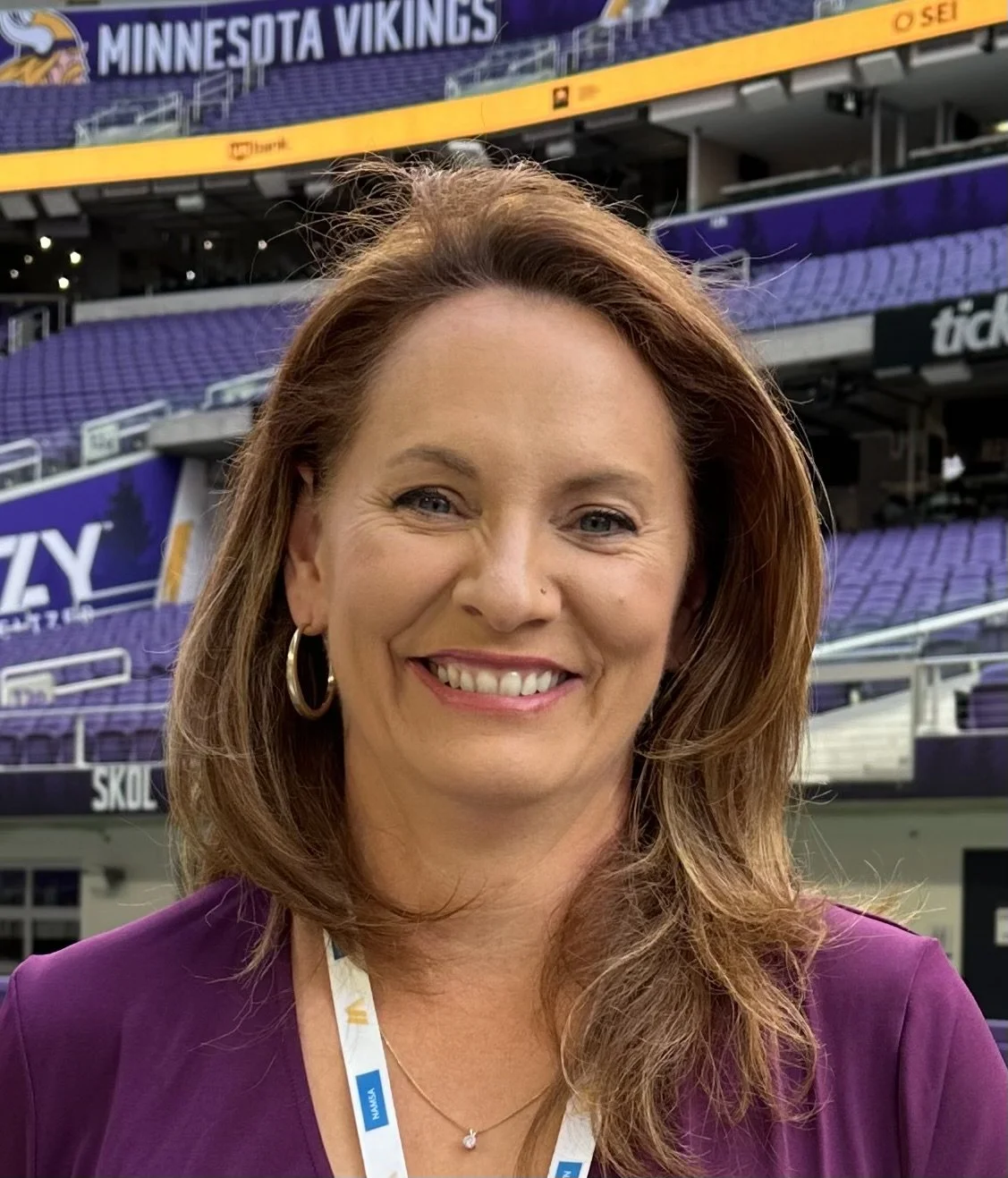 A smiling woman with red hair and hoop earrings standing in a stadium with purple seats and signs that read 'Minnesota Vikings.'