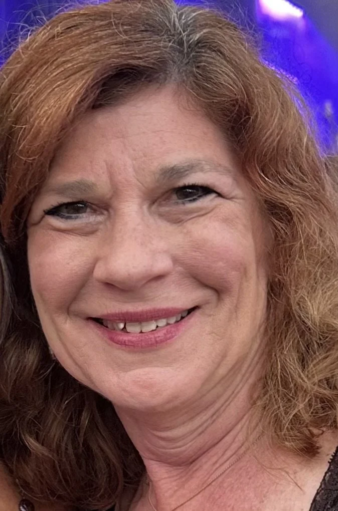 Close-up of a middle-aged woman with curly red hair smiling at the camera. Background features blue and purple lighting.