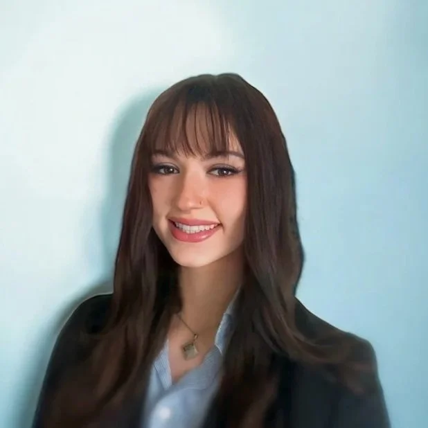 A young woman with long dark brown hair and bangs, smiling, wearing a black blazer over a light blue shirt, standing against a light blue background.