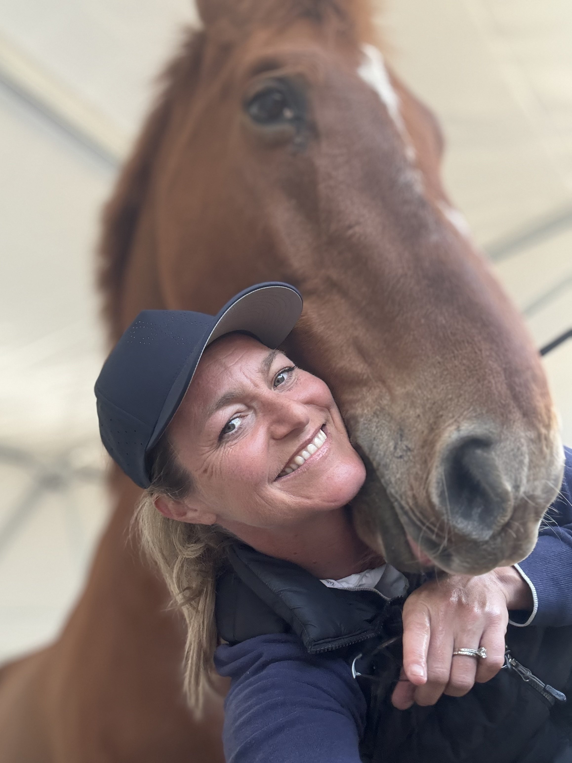 A woman smiling with a horse on her shoulder inside a stable.