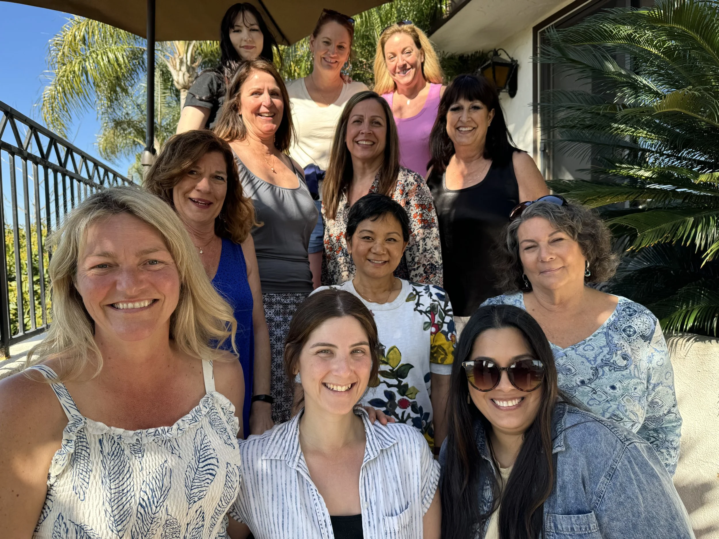 A group of fifteen women smiling outdoors under a large umbrella, with palm trees and a building in the background.