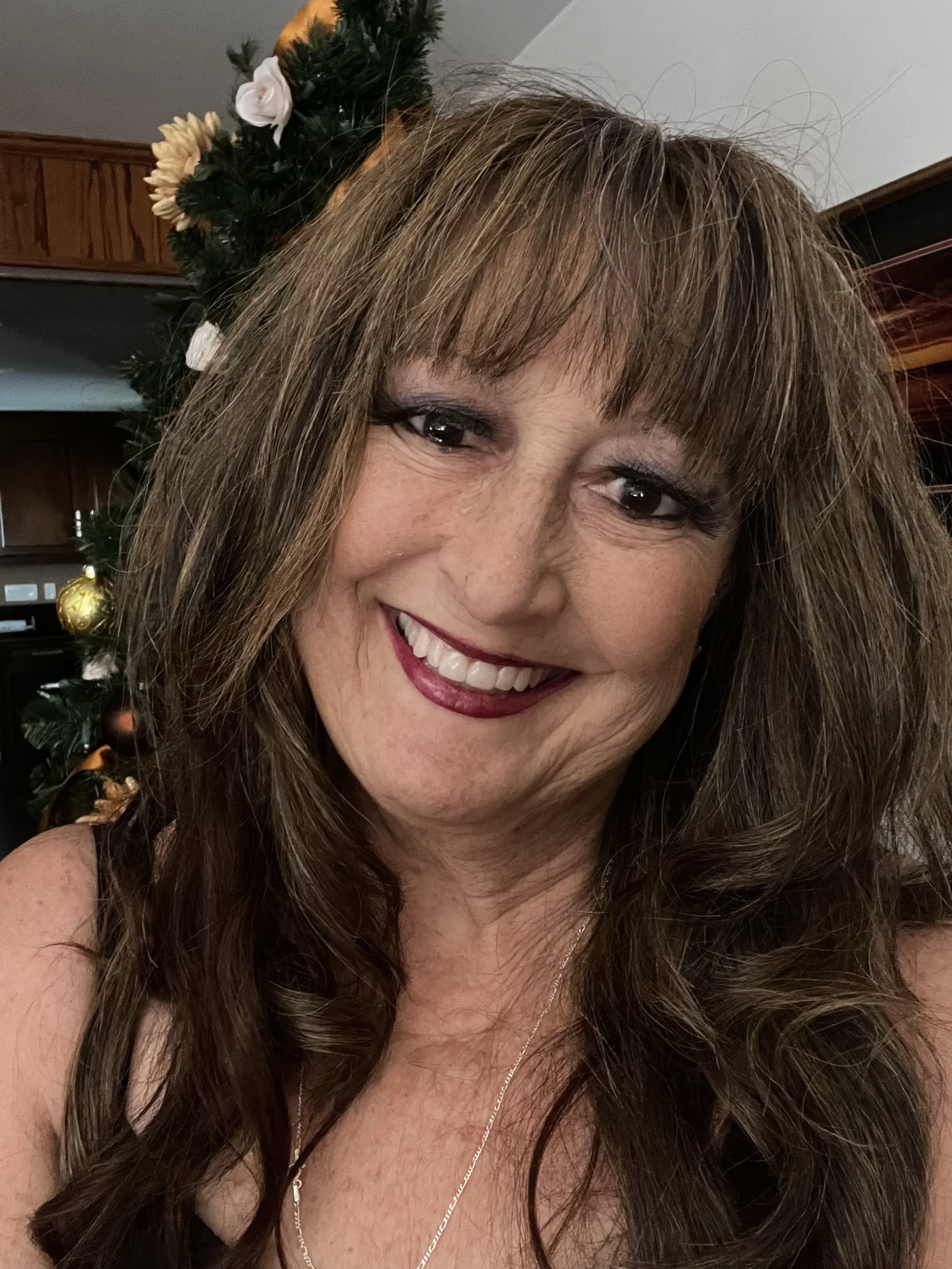 A woman with long brown hair and bangs smiling in front of a decorated Christmas tree.