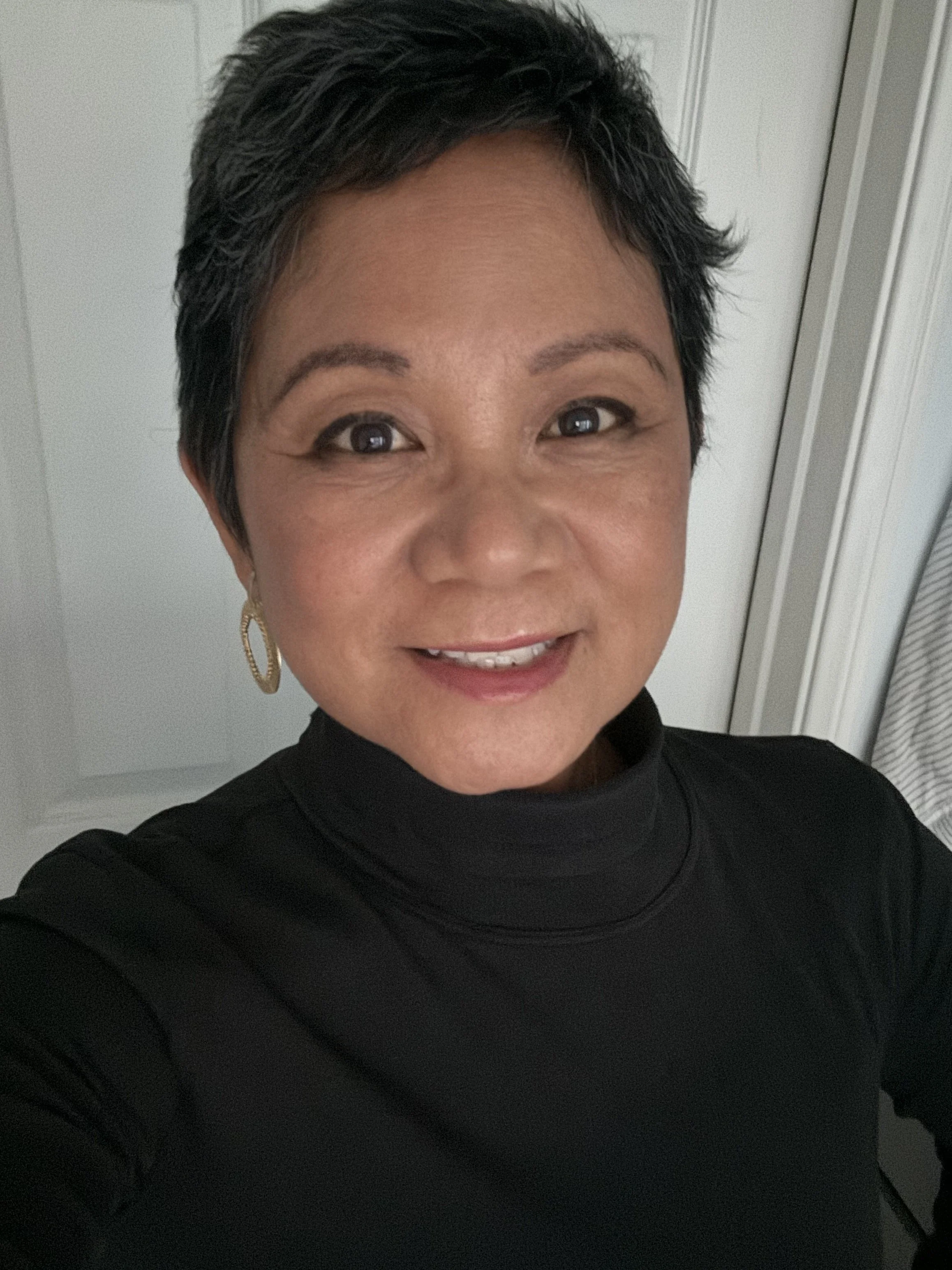 A woman with short black hair smiling at the camera, wearing a black turtleneck and gold hoop earrings, standing indoors near a white door.