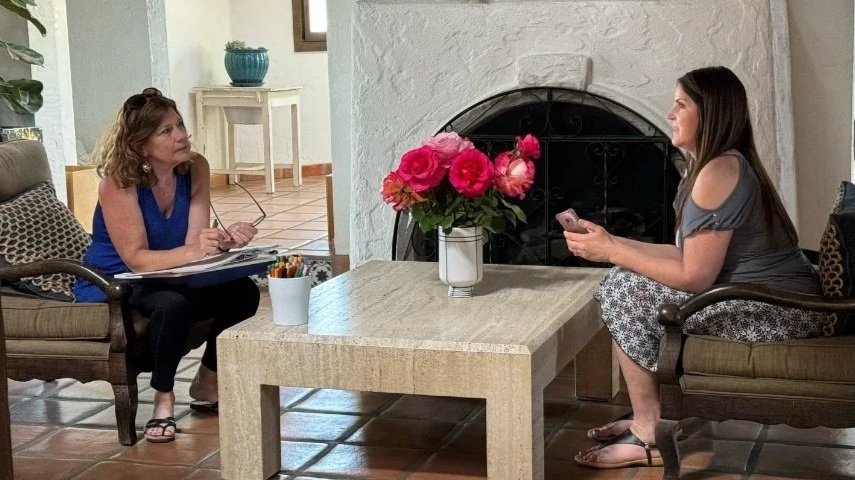 Two women sitting across from each other in a living room, engaging in conversation. One woman is taking notes, and the other woman is holding a phone. A coffee table with a vase of pink flowers is between them.