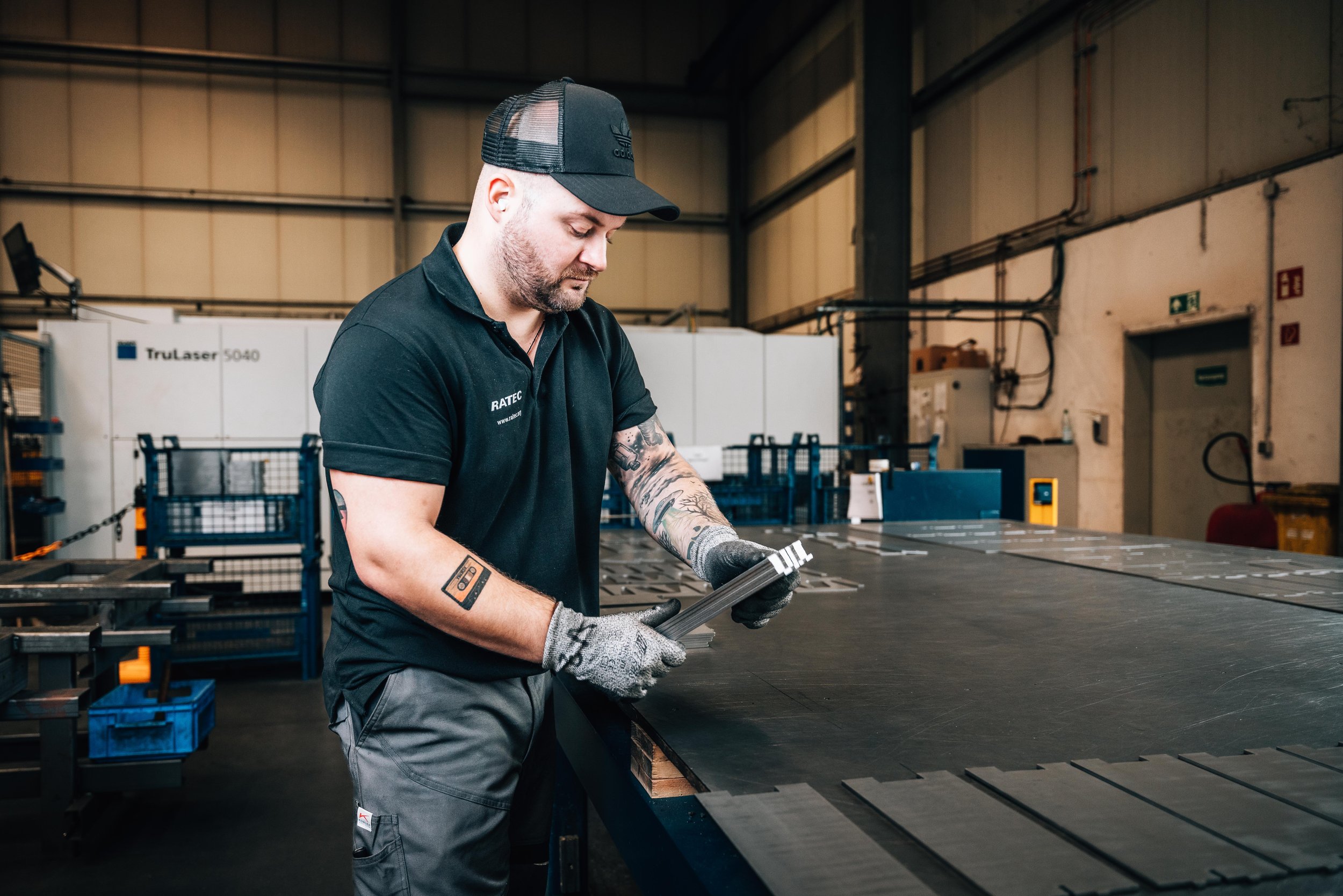 A man in a factory setting wearing a black polo shirt and cap works with metal sheets on a table. A TruLaser 5040 machine is in the background, and the man is wearing gloves.