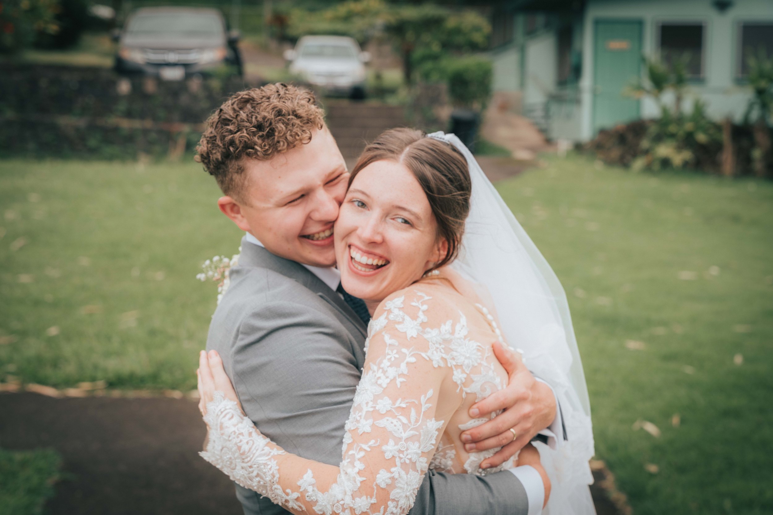 Bride and groom hugging and smiling outdoors, wearing wedding attire.
