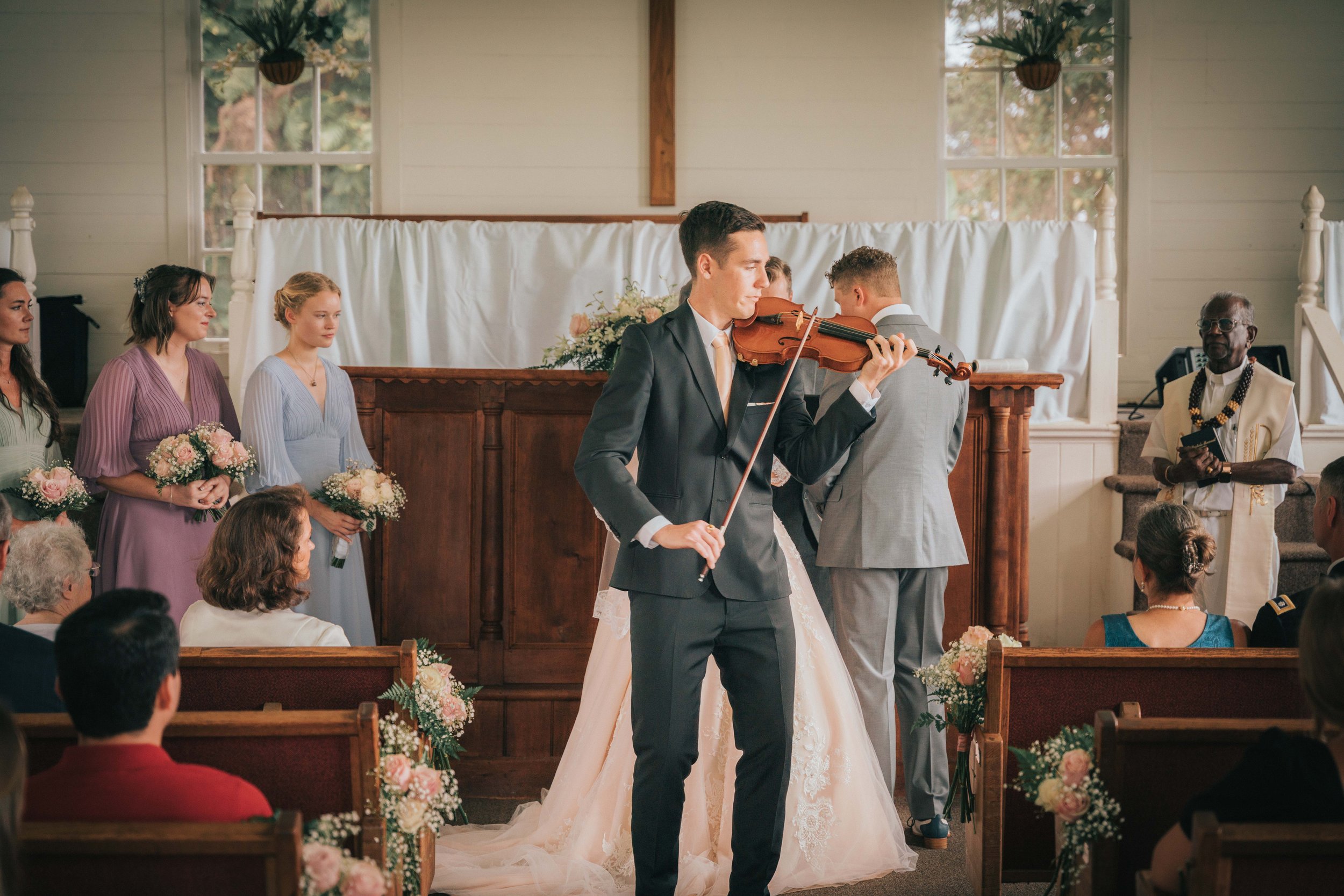 Indoor wedding ceremony with a violinist in a suit playing while standing in front of the altar. Bridesmaids in pastel dresses and bouquets are on the left, and a couple is in the center, partially visible. An officiant, dressed in white with a stole