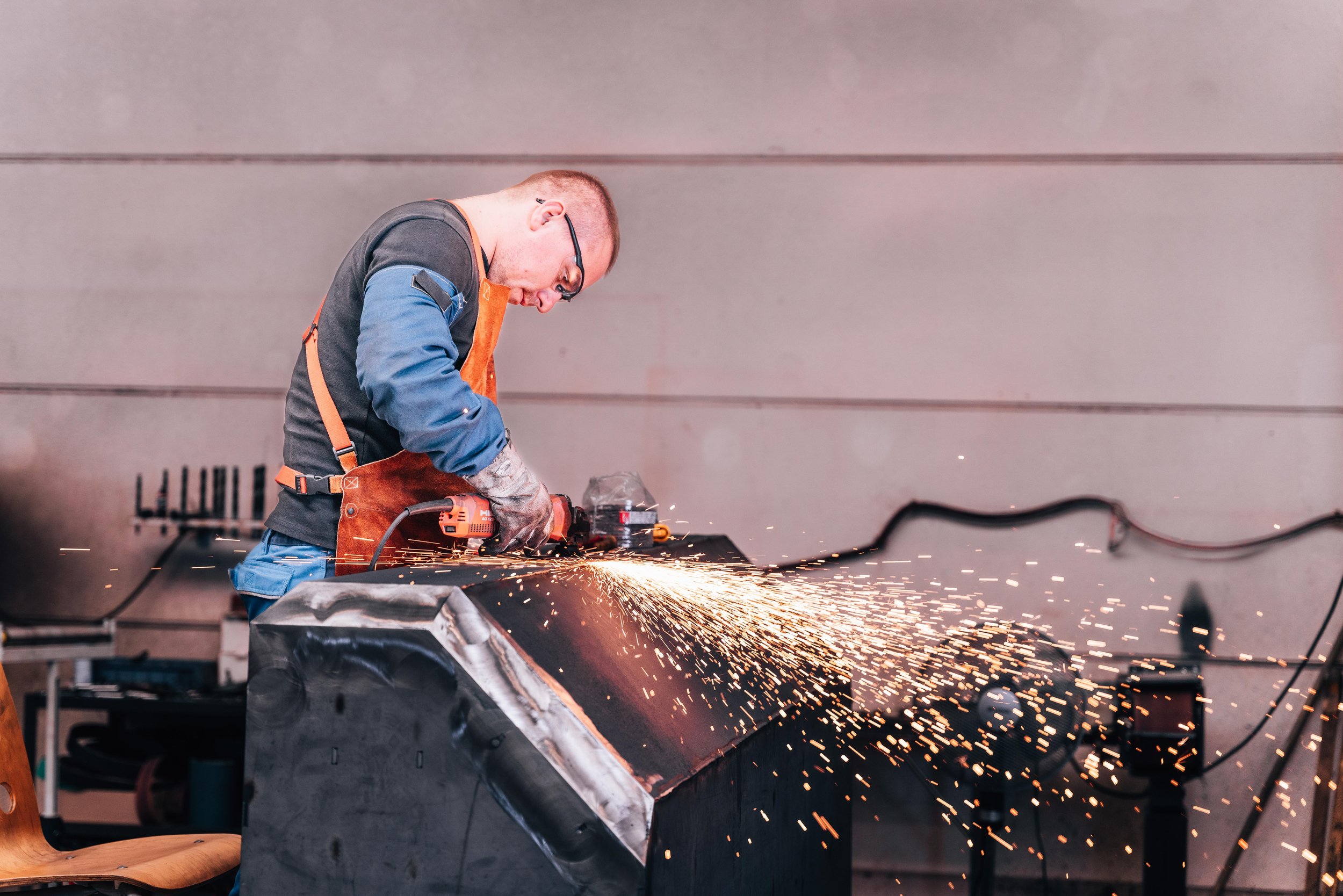 Man wearing safety goggles and apron using an angle grinder with sparks flying in a workshop.