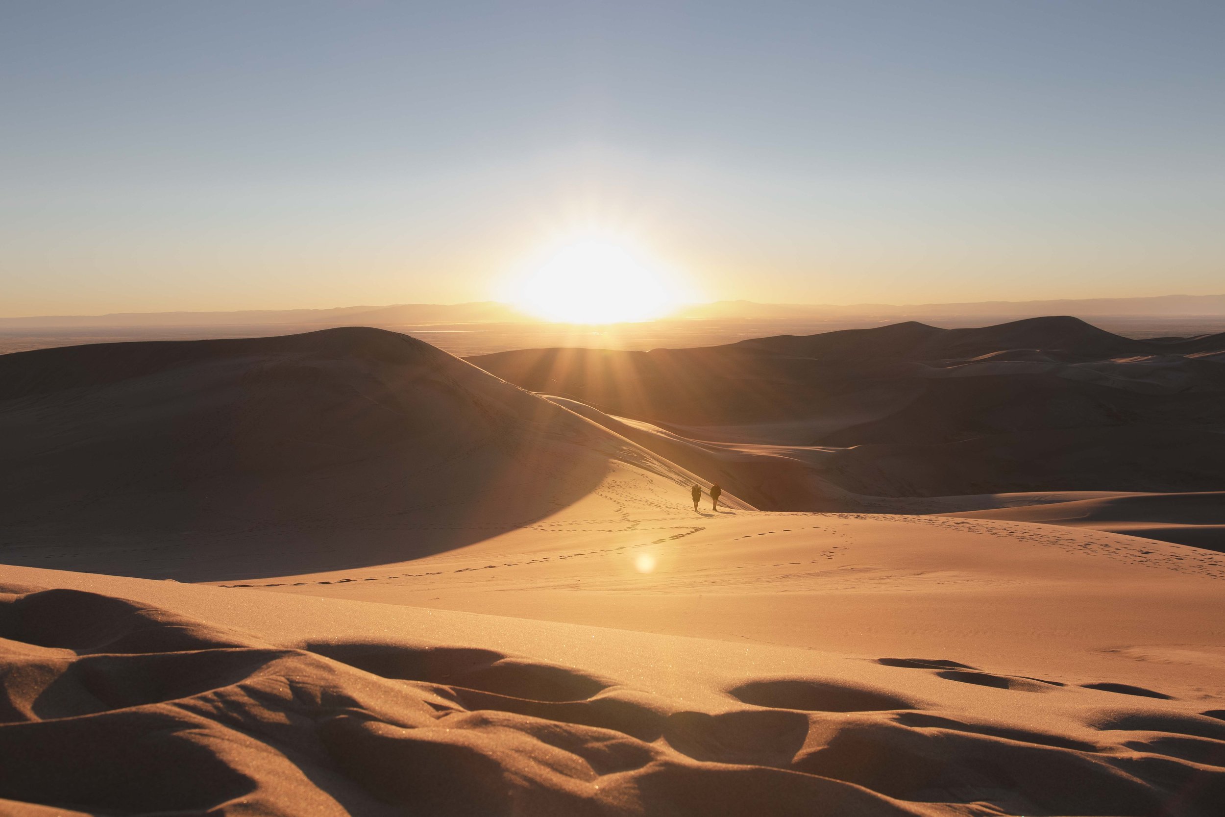 Sunset over sand dunes with footprints and two people walking.