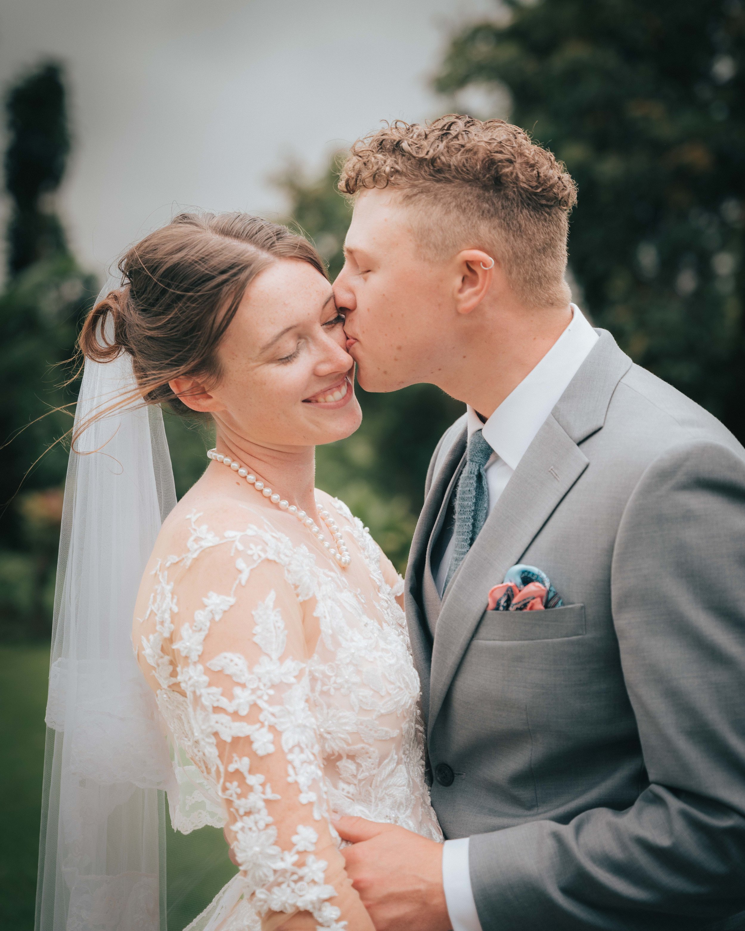 Bride and groom standing outdoors, bride in lace dress and veil, groom in gray suit, sharing a tender moment. The groom is kissing the bride's cheek.