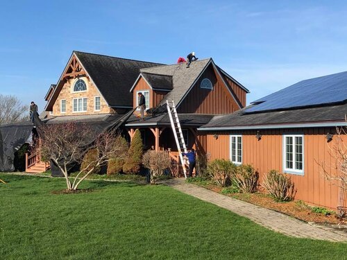 Workers installing or repairing shingles on a multi-story house with a steep roof, surrounded by a well-maintained lawn and garden, on a clear sunny day.