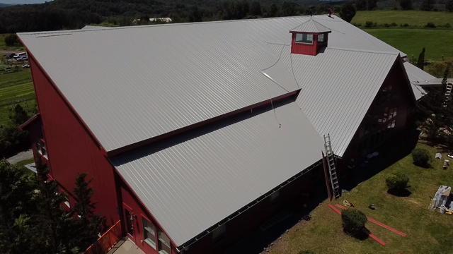 Aerial view of a large red barn with a silver metal roof, a small cupola, and a ladder leaning against the side, surrounded by green fields and trees.