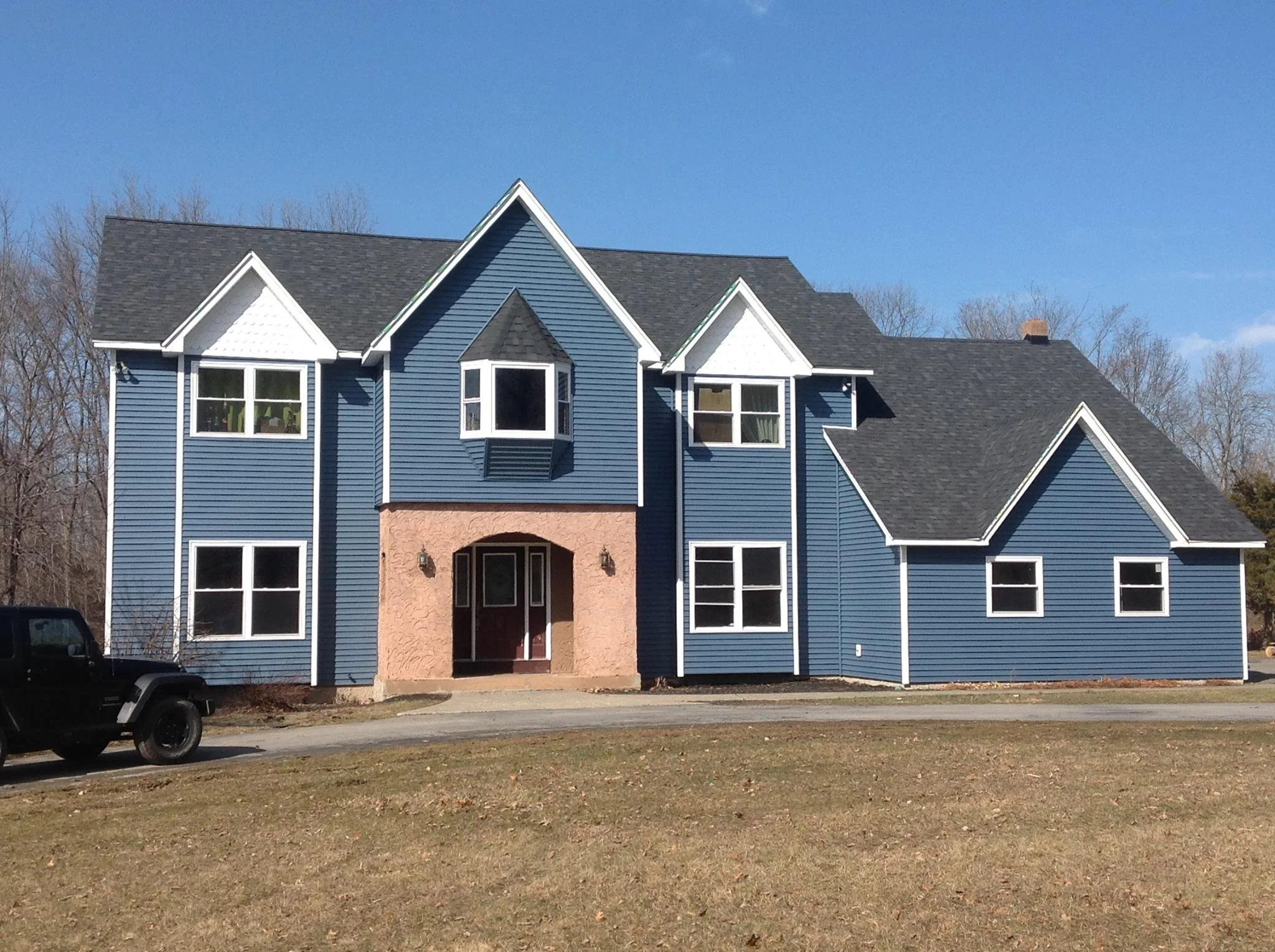 A large two-story house with blue vinyl siding, white trim, and a gray shingle roof. The house features multiple gables, several double-hung windows, and a front porch with an archway. There is a black SUV parked on the street in front of the house.
